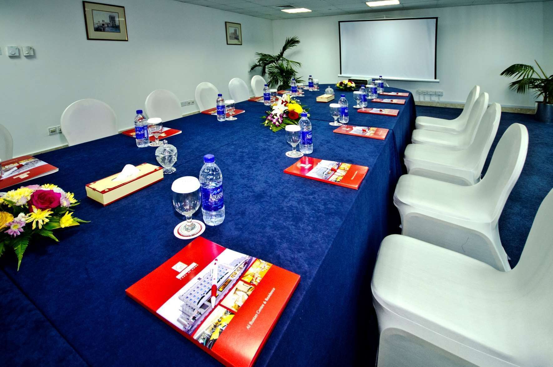 Uma sala de conferência com uma mesa comprida, cadeiras brancas, garrafas de água, copos e pastas vermelhas.