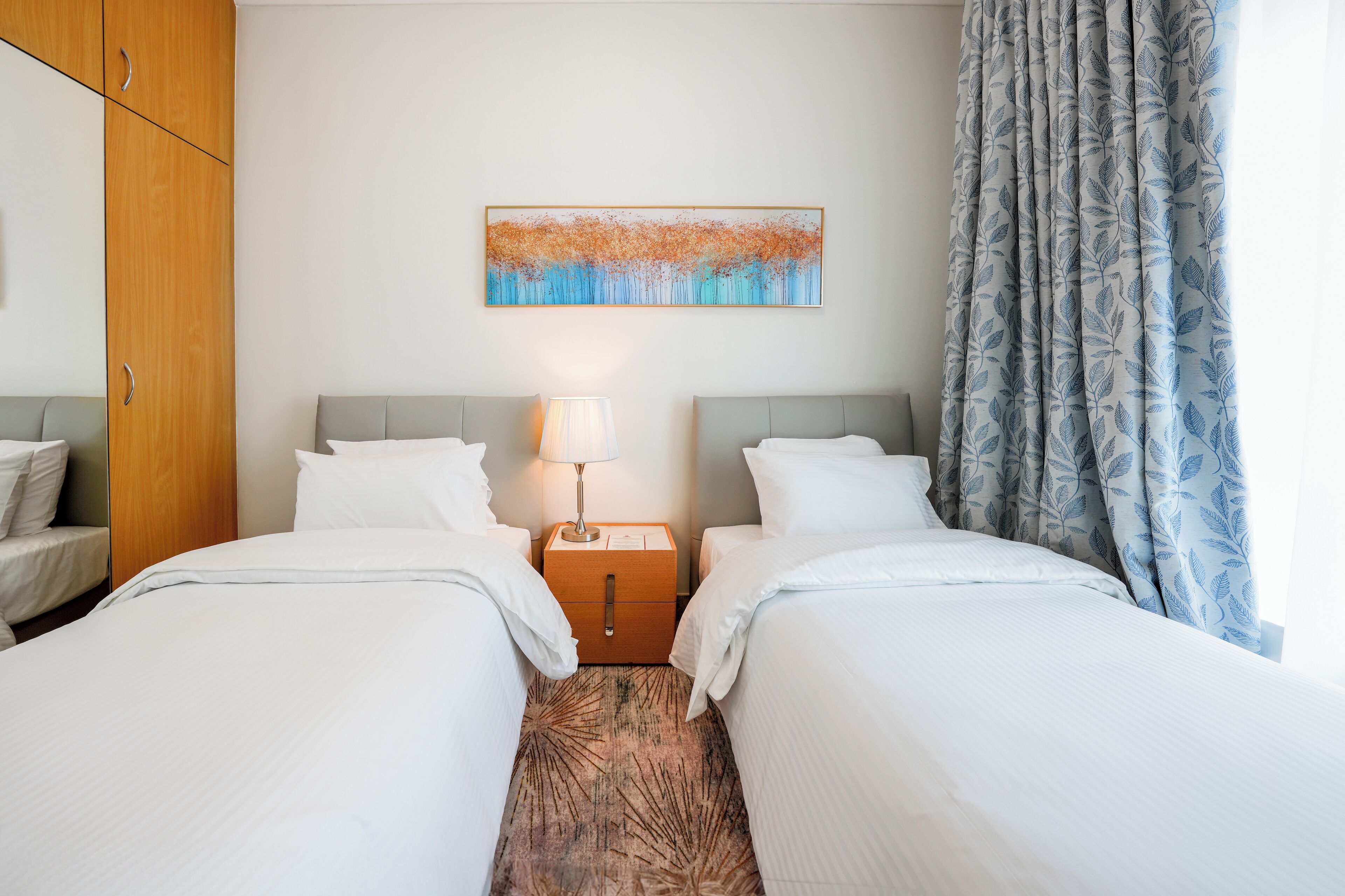 A hotel room with two beds, a nightstand with a lamp, a wooden wardrobe, and a painting on the wall.