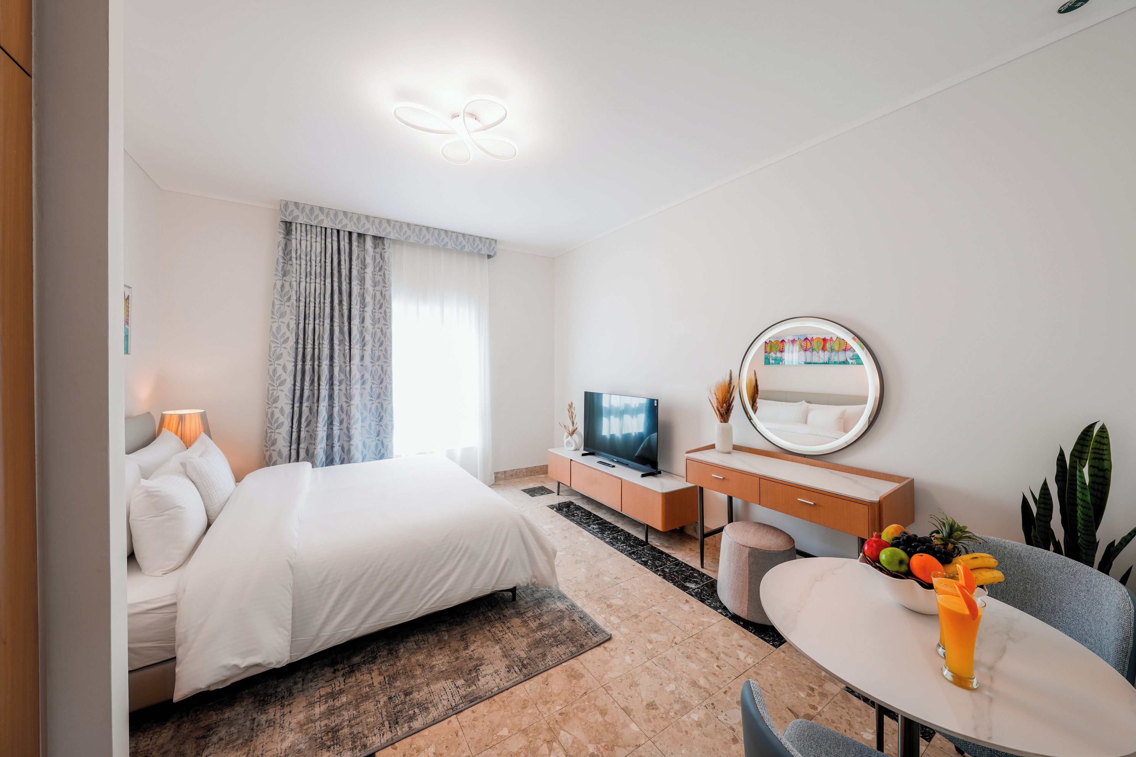 Ein modernes Hotelzimmer mit Bett, Schreibtisch, Fernseher und einer Essecke mit Obstschale und Glas Saft.