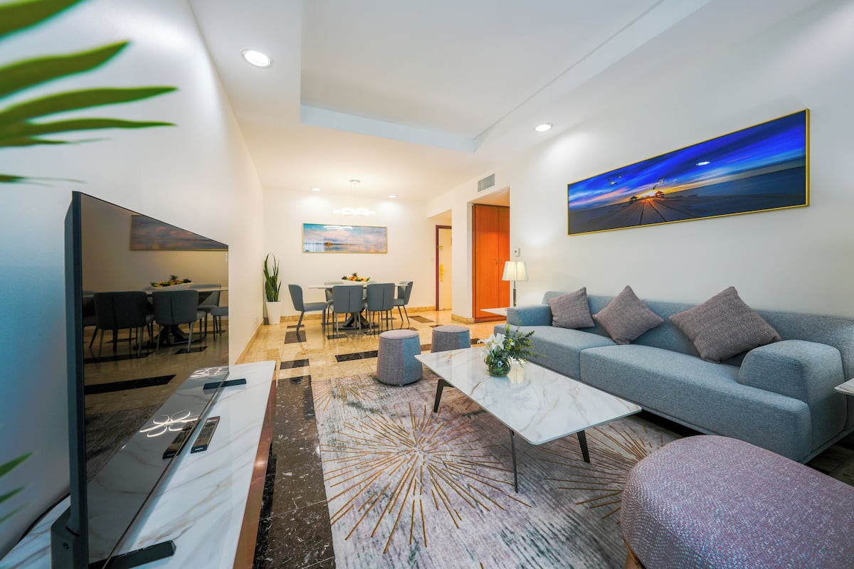 Un salon moderne avec un canapé, une table basse et un téléviseur. On y trouve également un coin repas avec des chaises et une table, ainsi qu’un tableau accroché au mur.