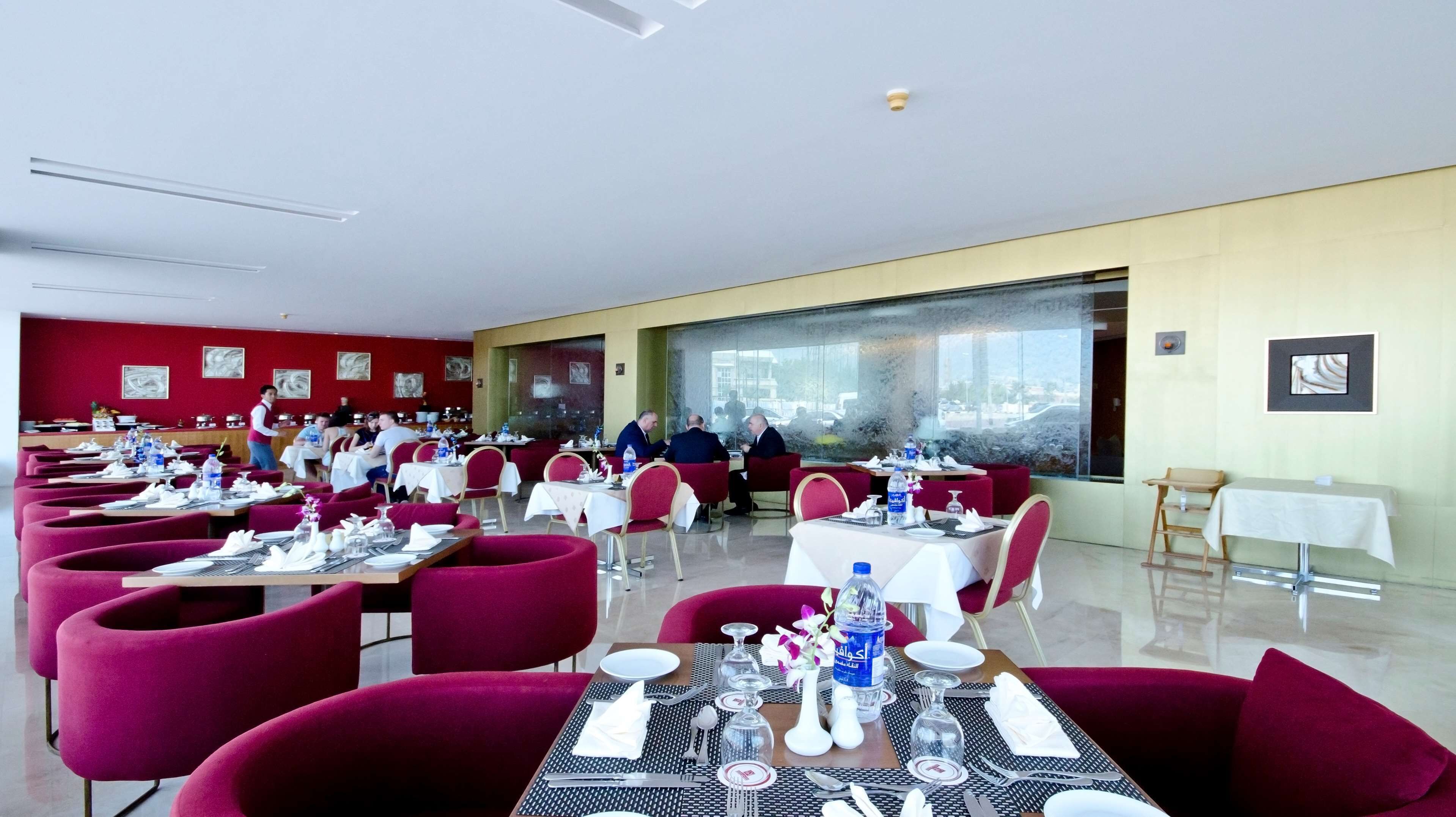 Ein Restaurant mit mehreren Tischen, roten Stühlen und Blick auf die Außenwelt durch große Fenster.