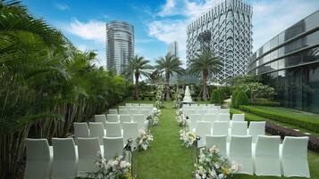 A wedding ceremony setup with white chairs and floral decorations, set against a backdrop of modern buildings and lush greenery.