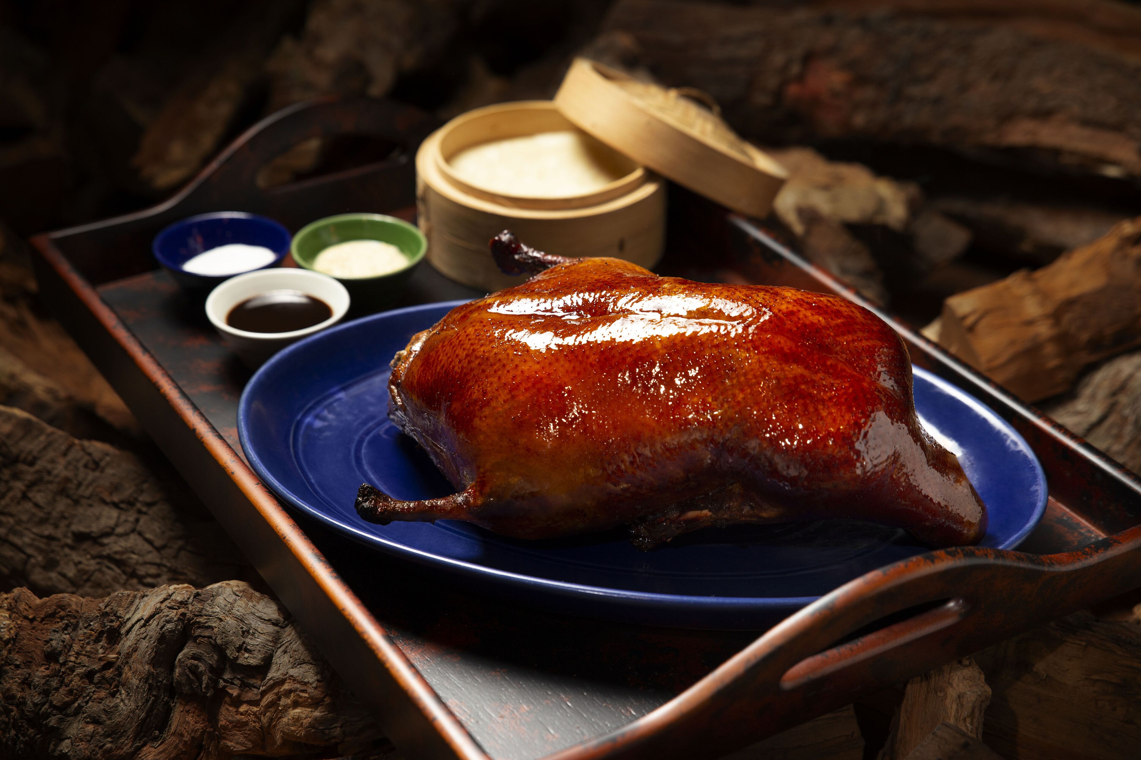 A roasted duck on a blue plate with a tray, accompanied by small bowls of sauces.