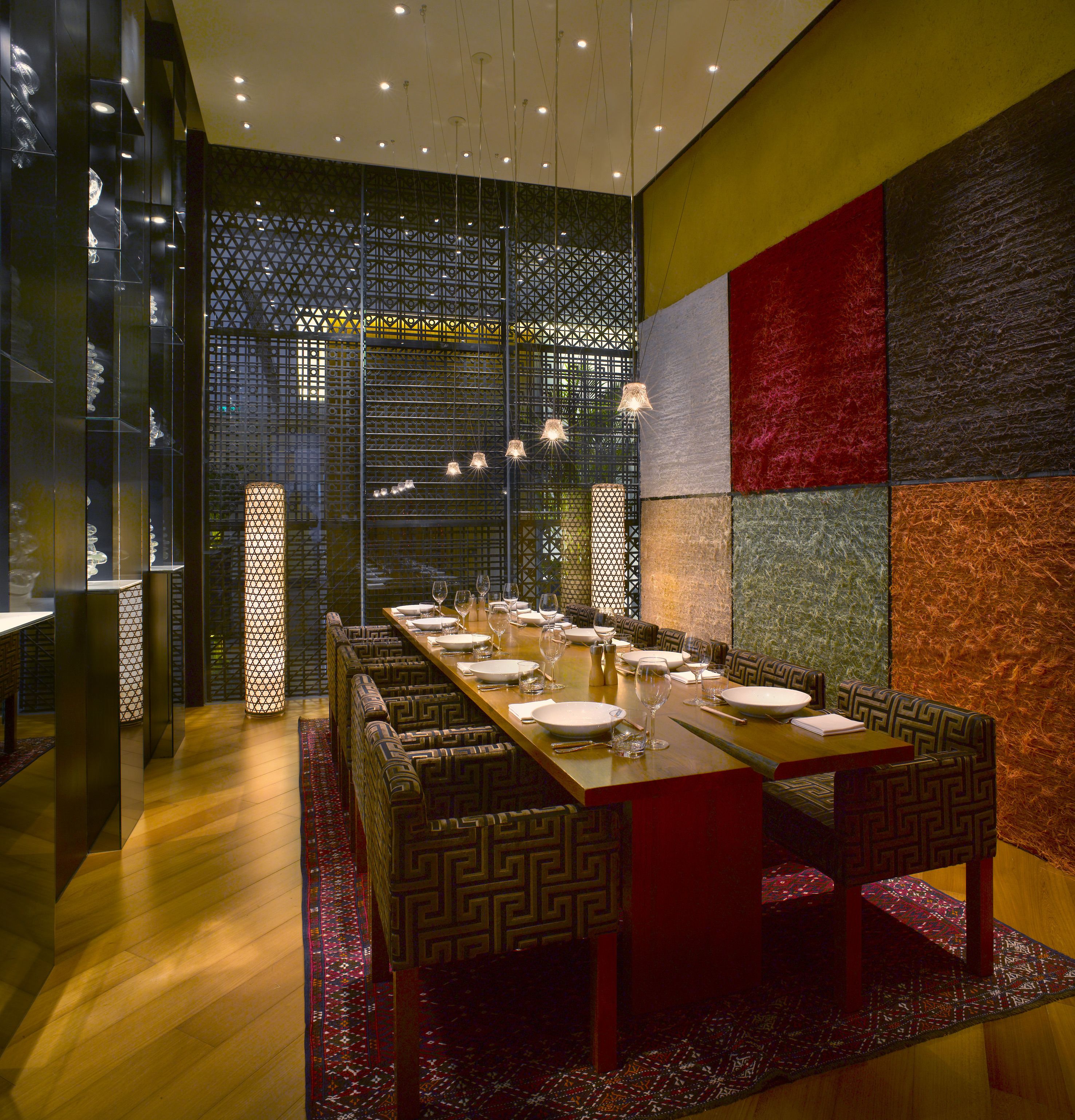Un comedor moderno con una larga mesa dispuesta para varias personas, bancos con motivos estampados y una pared decorativa con diseños geométricos.