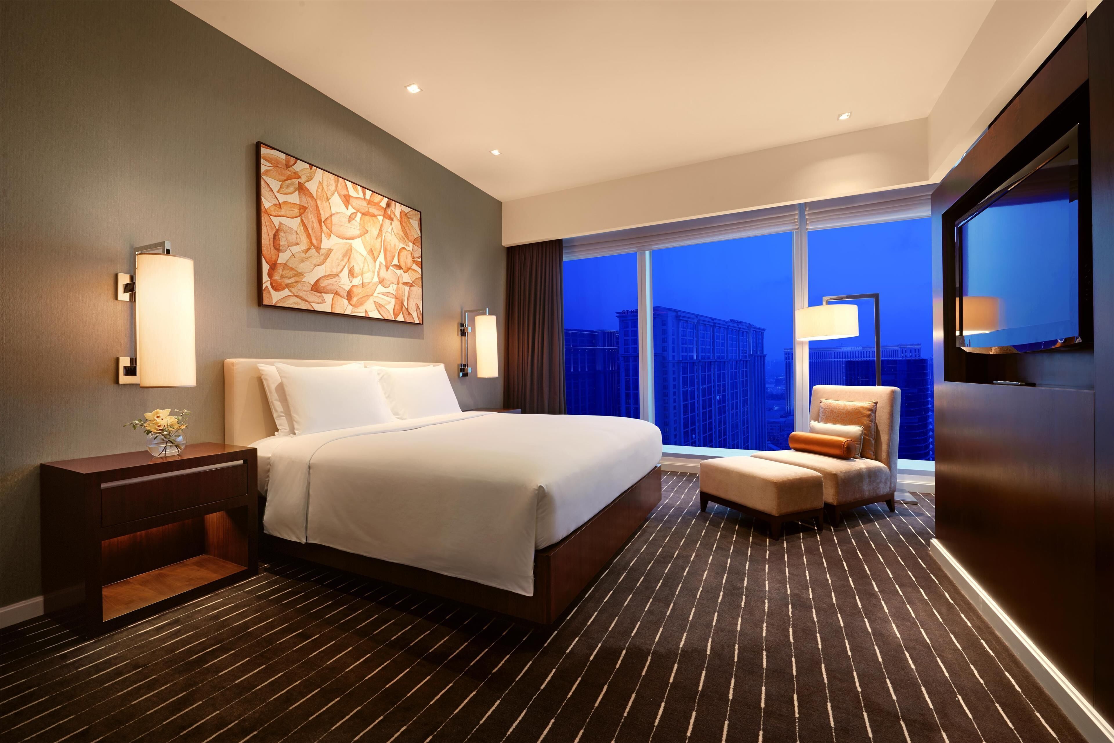 A modern hotel room with a large bed, a seating area, a large window with a city view, and a painting on the wall.
