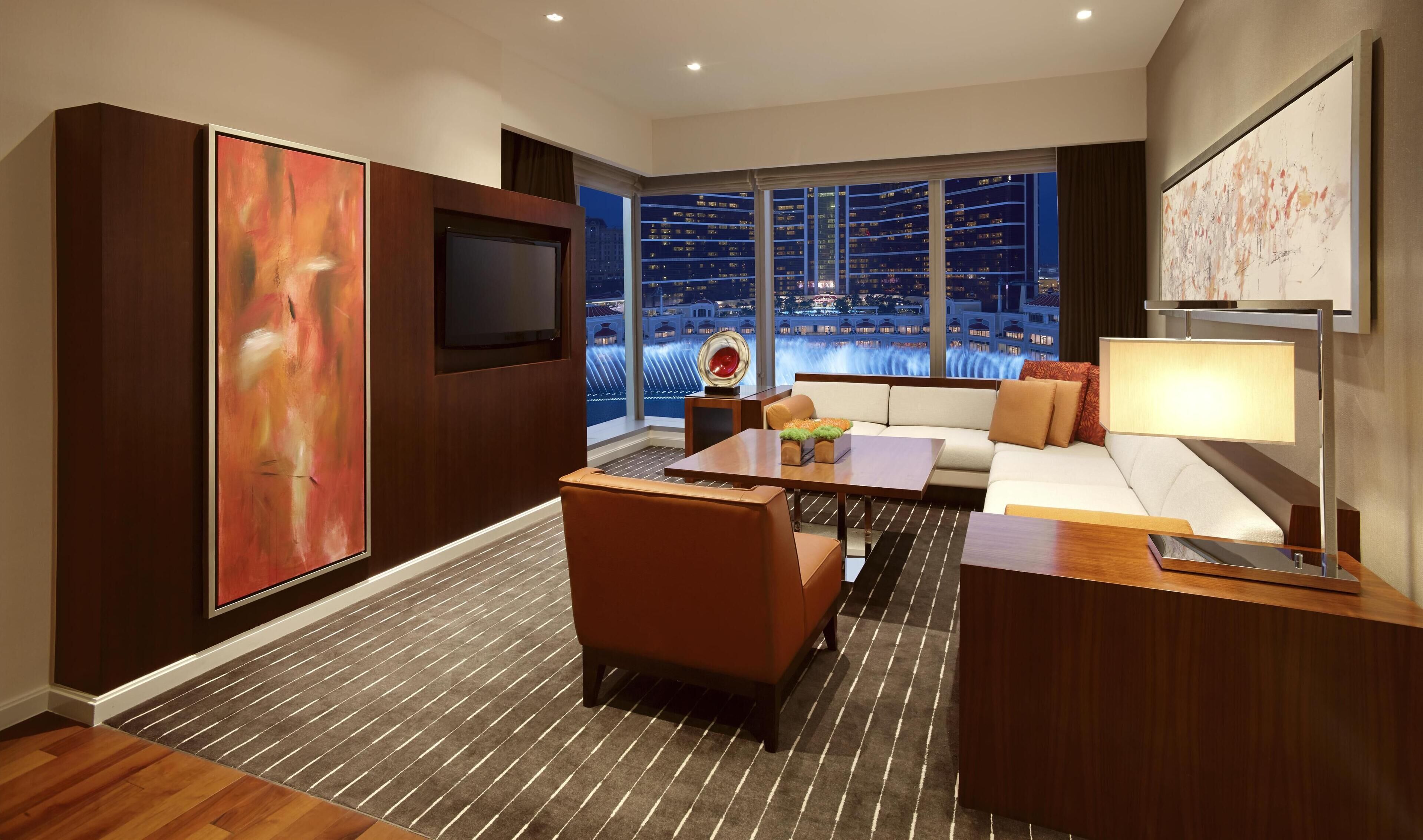 A modern hotel room with a large window offering a city view, a flat-screen TV, a sofa, a coffee table, and a large abstract painting.