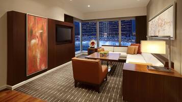 A modern hotel room with a large window offering a city view, a flat-screen TV, a sofa, a coffee table, and a large abstract painting.