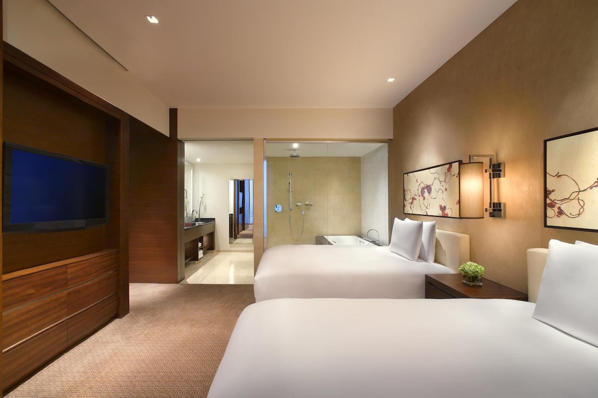 Habitación de hotel con dos camas, televisión, baño con ducha y un pasillo que conduce a otra habitación.