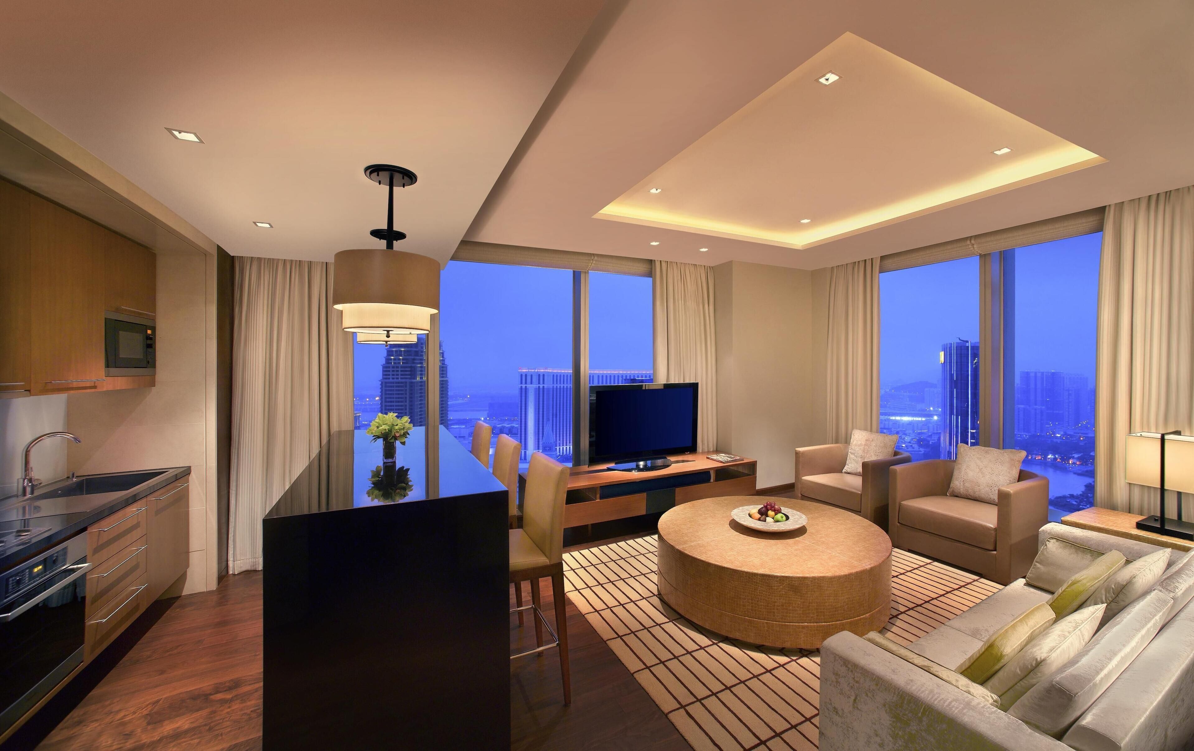 A modern hotel room with a kitchen, a dining area, a living room with a sofa and armchairs, a TV, and large windows offering city views.