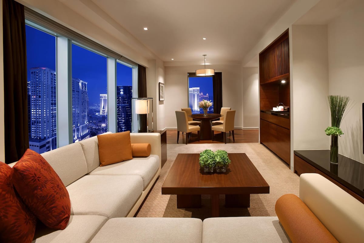 Una habitación de hotel moderna con un gran ventanal que ofrece vistas a la ciudad, una zona de estar acogedora con sofá y sillones, una mesa de comedor con sillas, y un televisor de pantalla plana montado en la pared.
