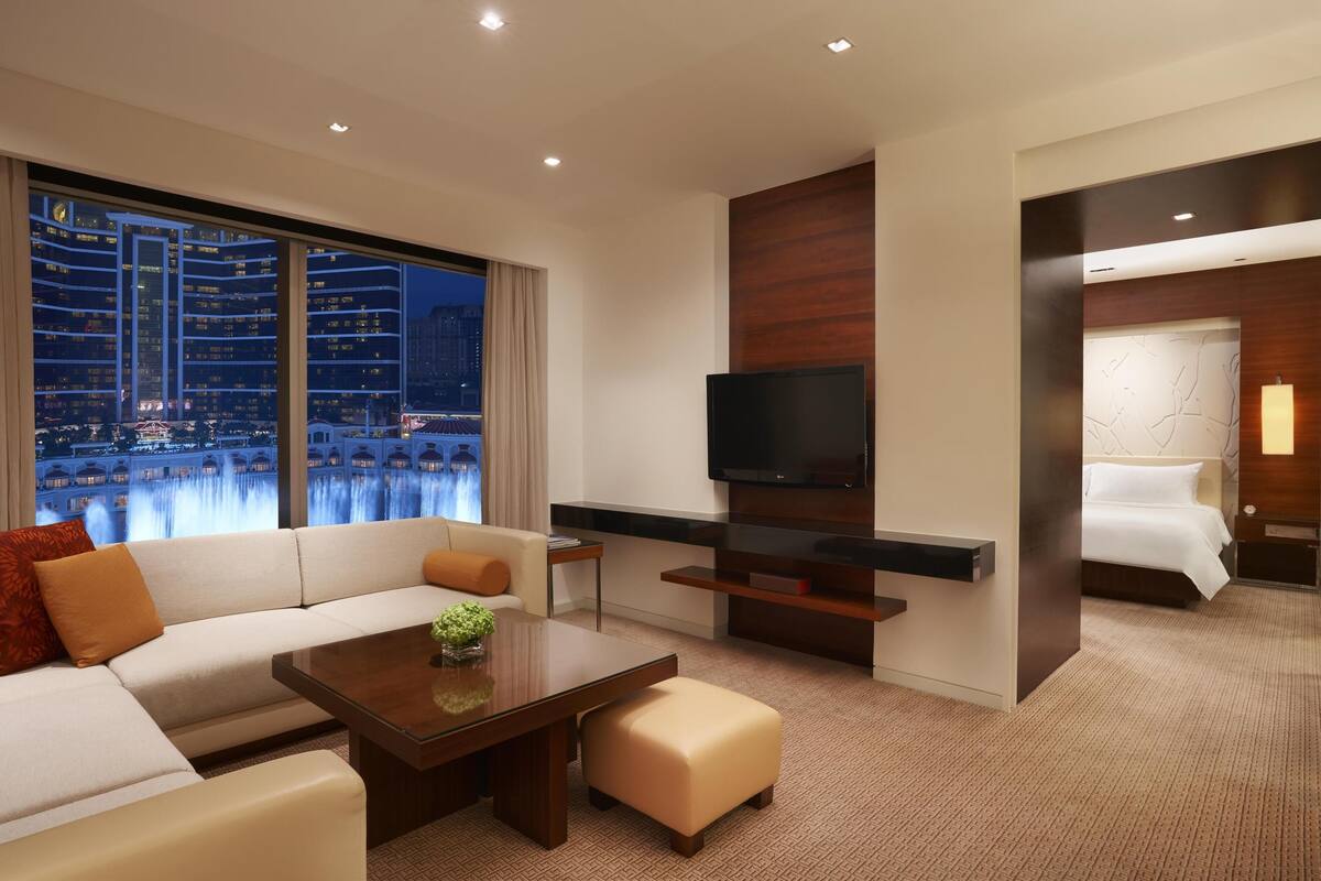 A modern hotel room with a large window offering a city view, a flat-screen TV, a sofa, a coffee table, and a separate bedroom area.