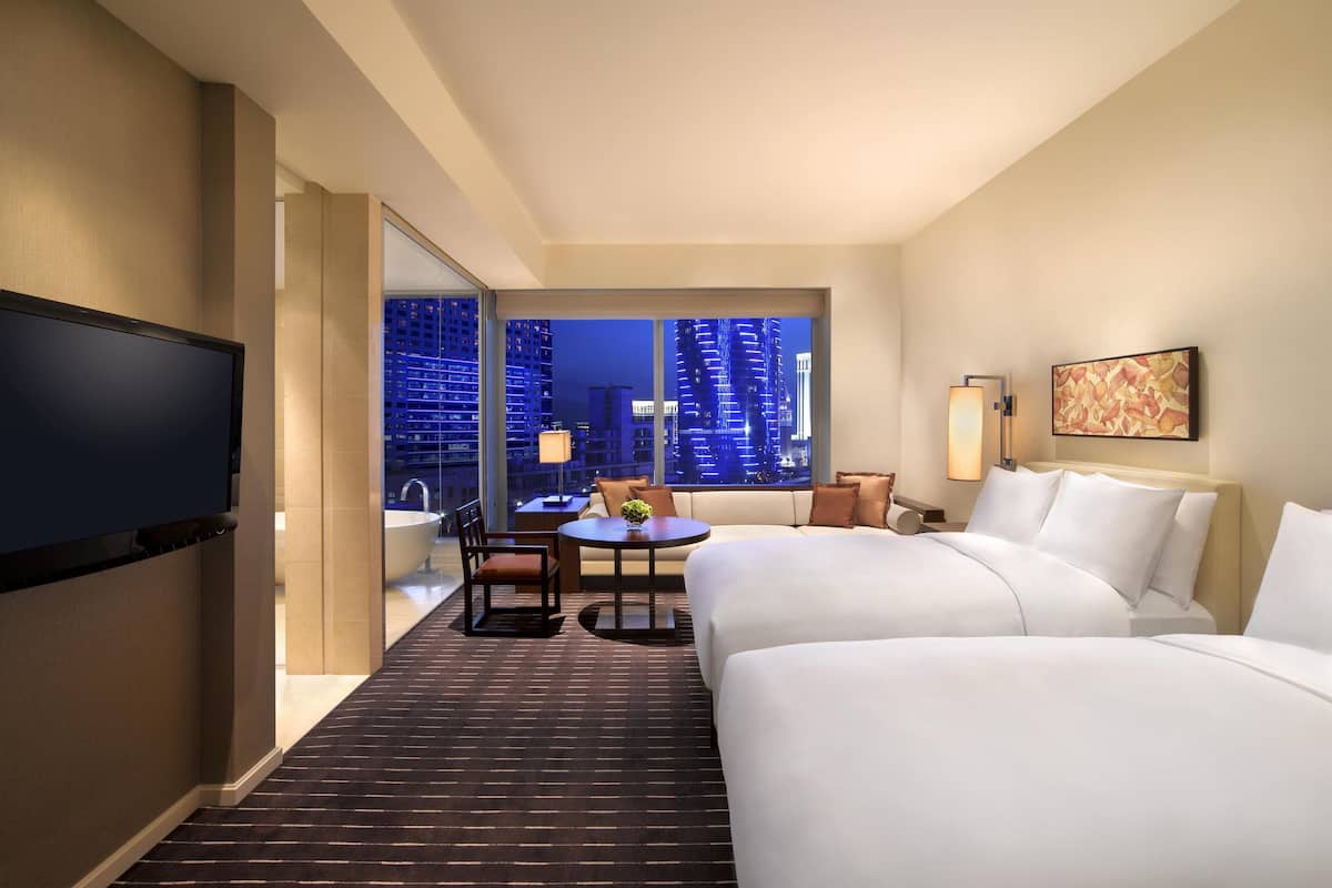 Habitación de hotel con una cama grande, televisión, una mesita con sillas y vista a la ciudad de noche.
