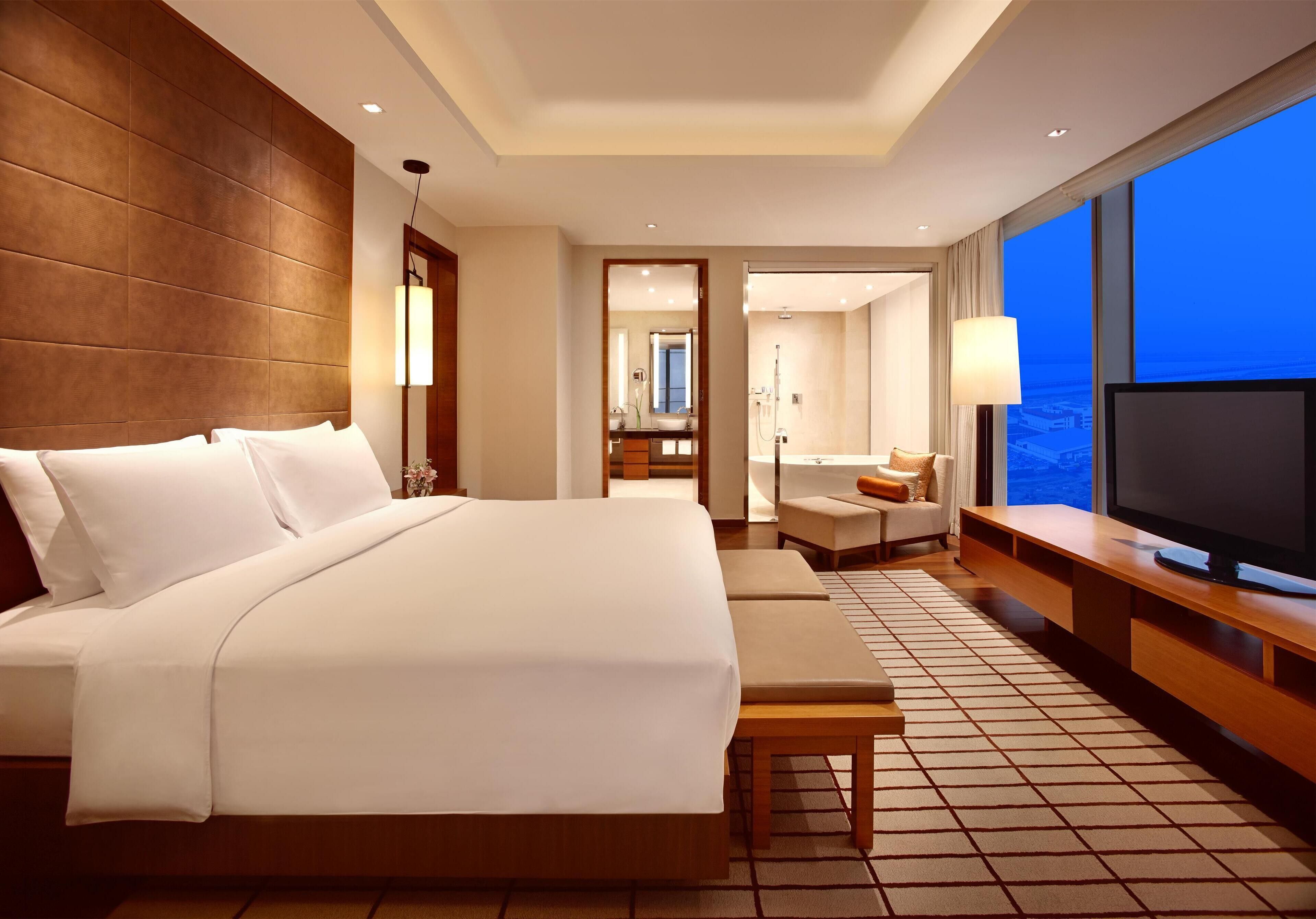 Habitación de hotel con una cama grande, televisión, zona de estar y vistas a la ciudad.