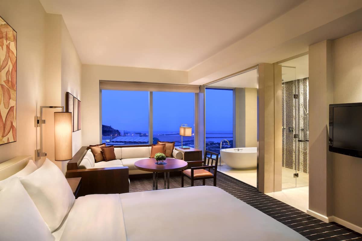 Habitación de hotel con una cama grande, una zona de estar con mesa y sillas, una bañera y vistas de la ciudad al atardecer.