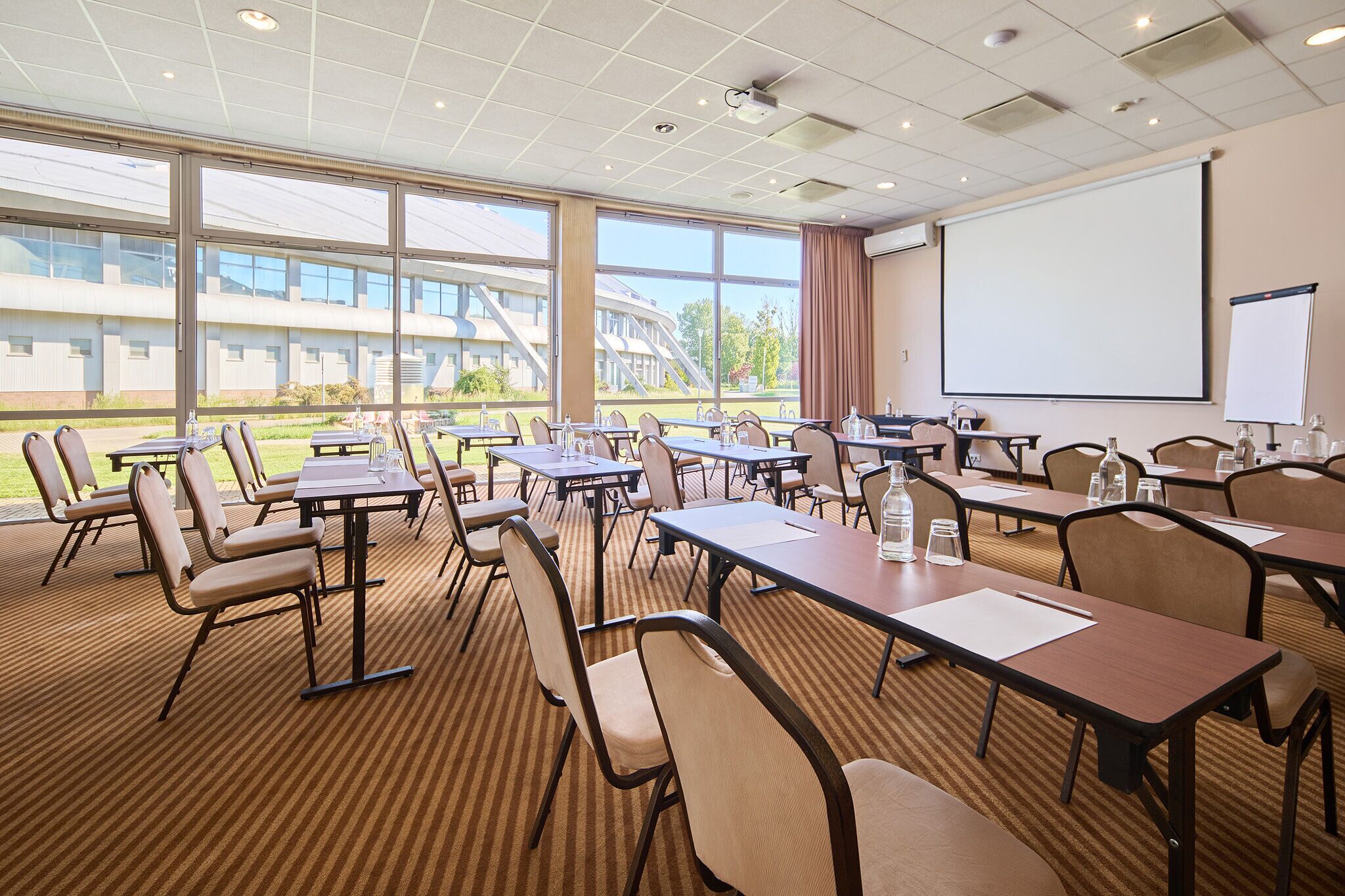 A conference room with tables and chairs, large windows, and a projector screen.