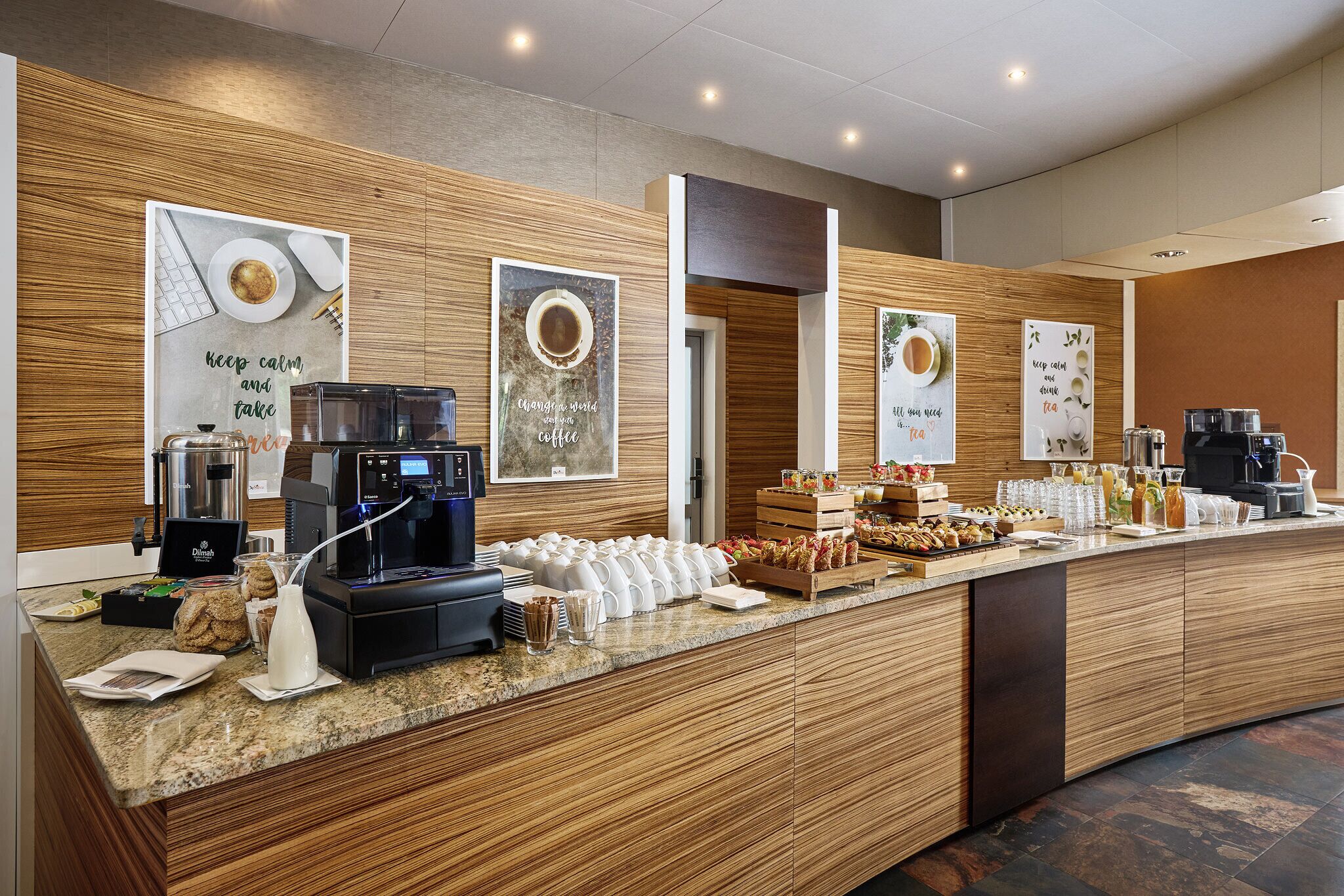 A hotel breakfast area with a coffee machine, tea station, and a variety of food items on the counter.