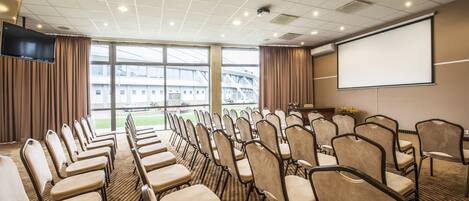 A conference room with rows of chairs, a projector screen, and a large window with a view of buildings.