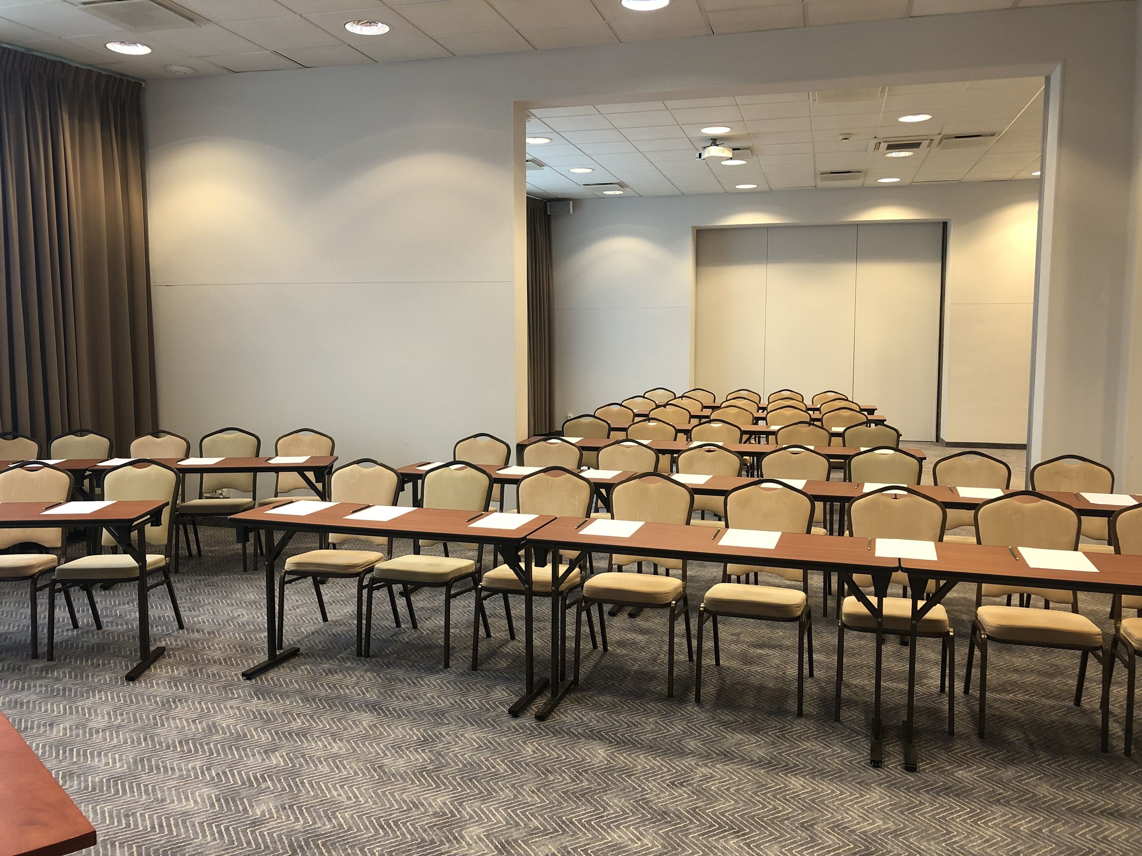 Un salón de conferencias con filas de sillas y mesas, una pantalla de proyector y un amplio ventanal con cortinas.