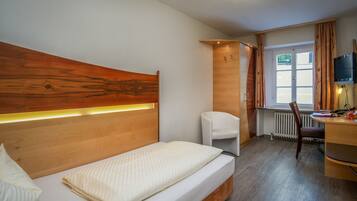 Habitación de hotel con cabecera de madera, una cama individual, un escritorio con silla, un radiador y una ventana con cortinas.