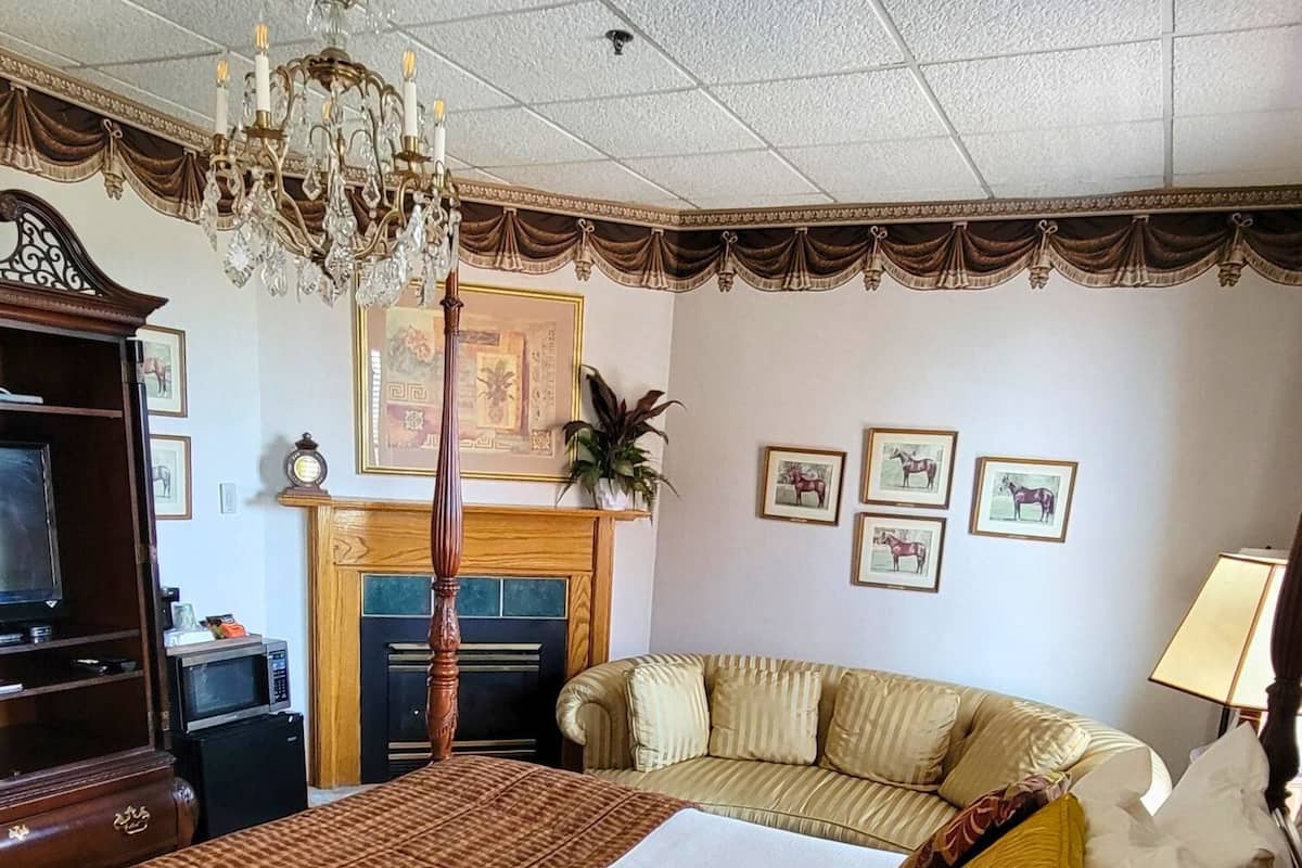 A hotel room with a large bed, a sofa, a chandelier, a fireplace, and framed pictures on the wall.