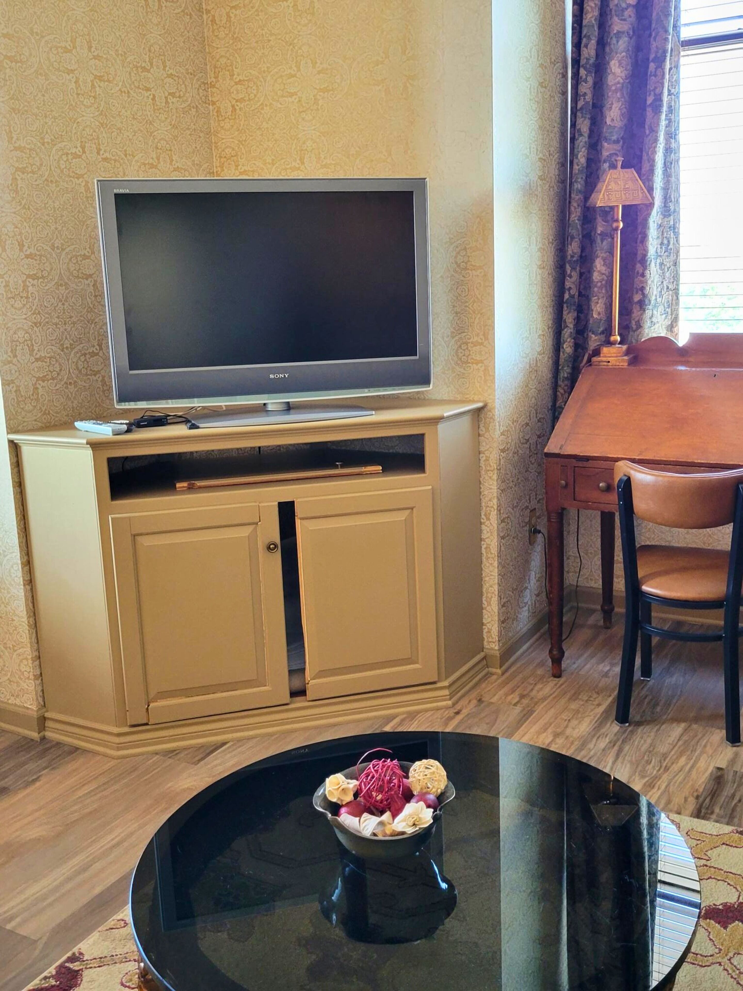 A television on a cabinet, a dining table with a bowl of fruit, and a lamp.