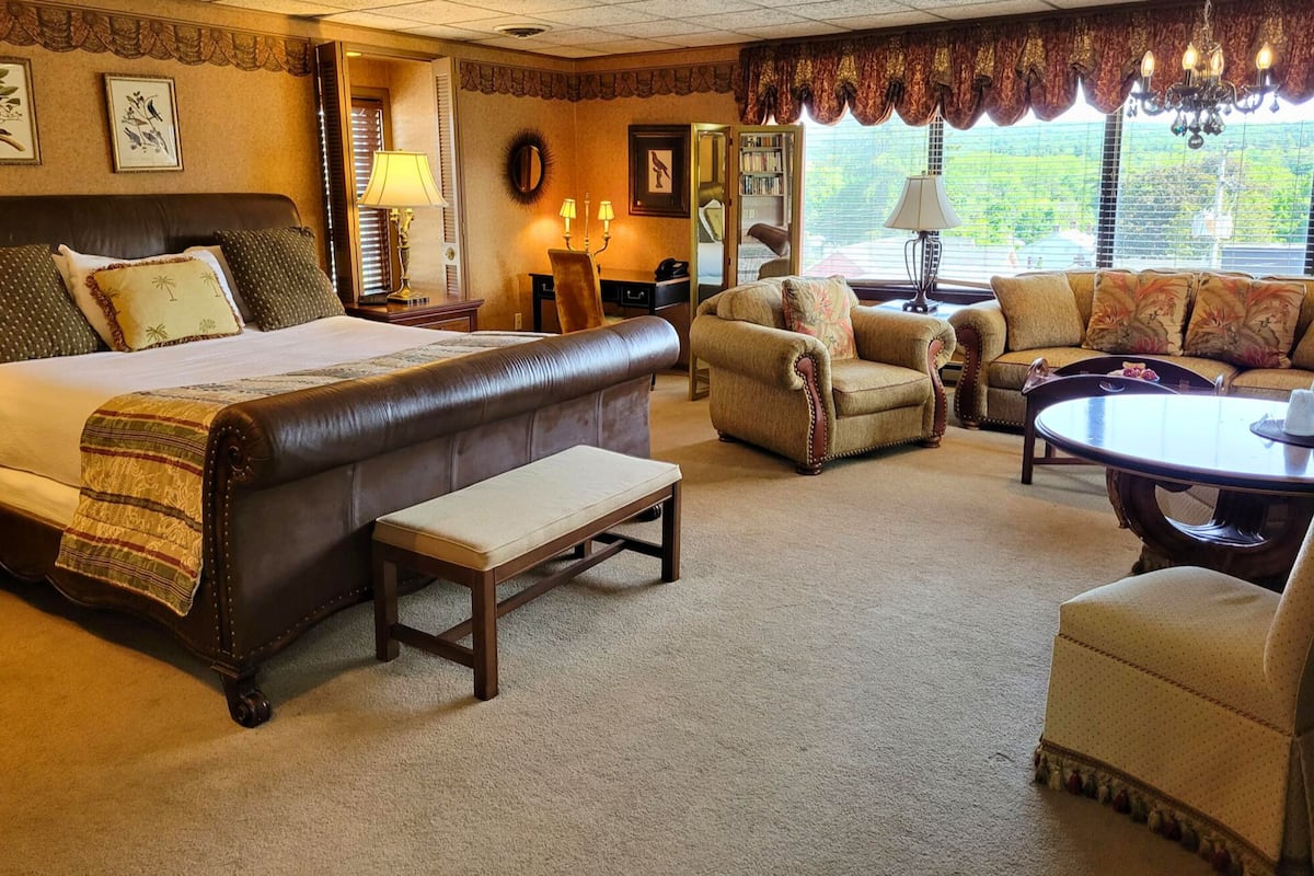 A spacious hotel room with a large bed, a sitting area with a sofa and armchairs, a small table, and a view of greenery through the windows.