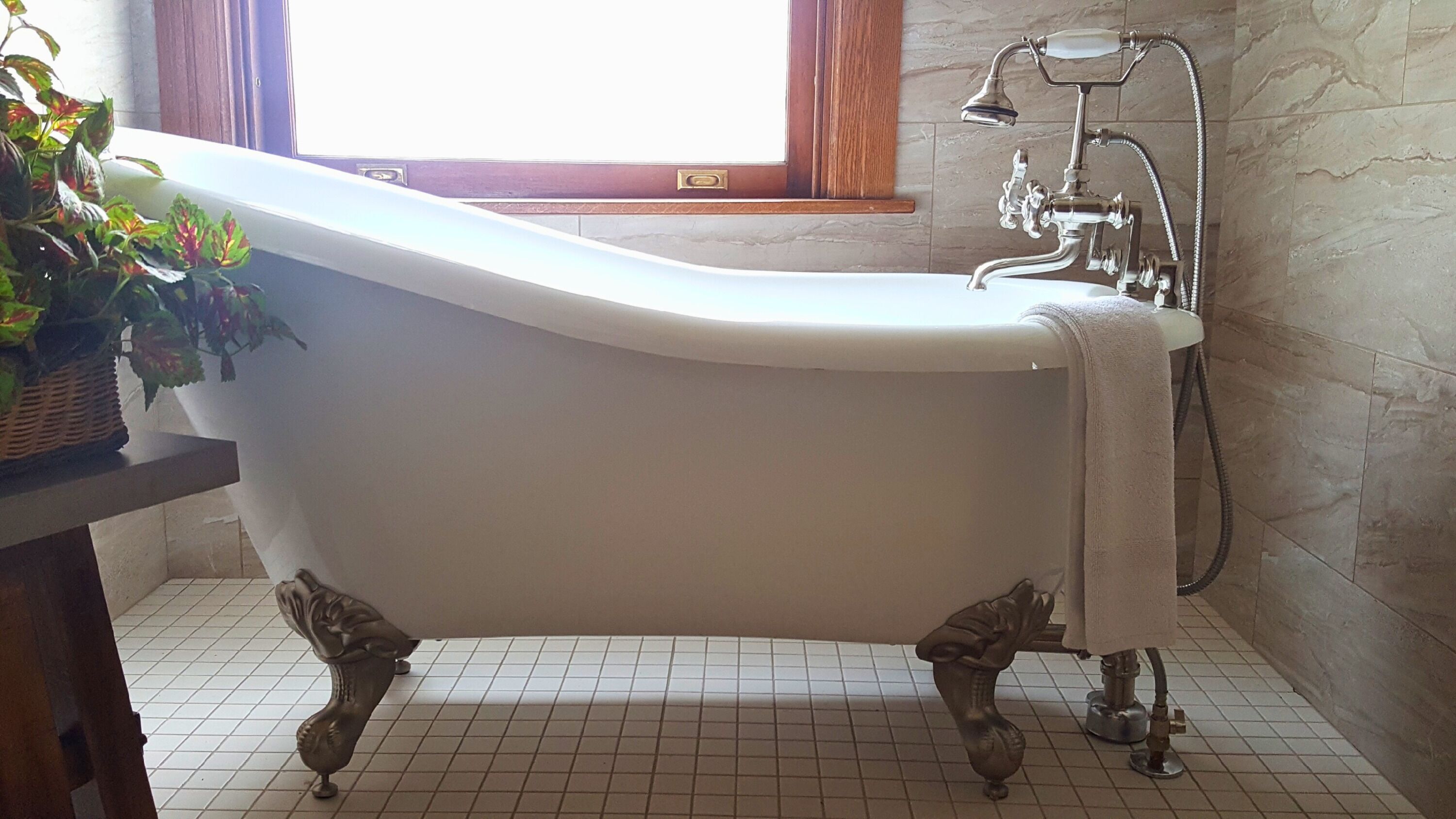A clawfoot bathtub with a shower attachment in a bathroom with tiled flooring and a window with curtains.