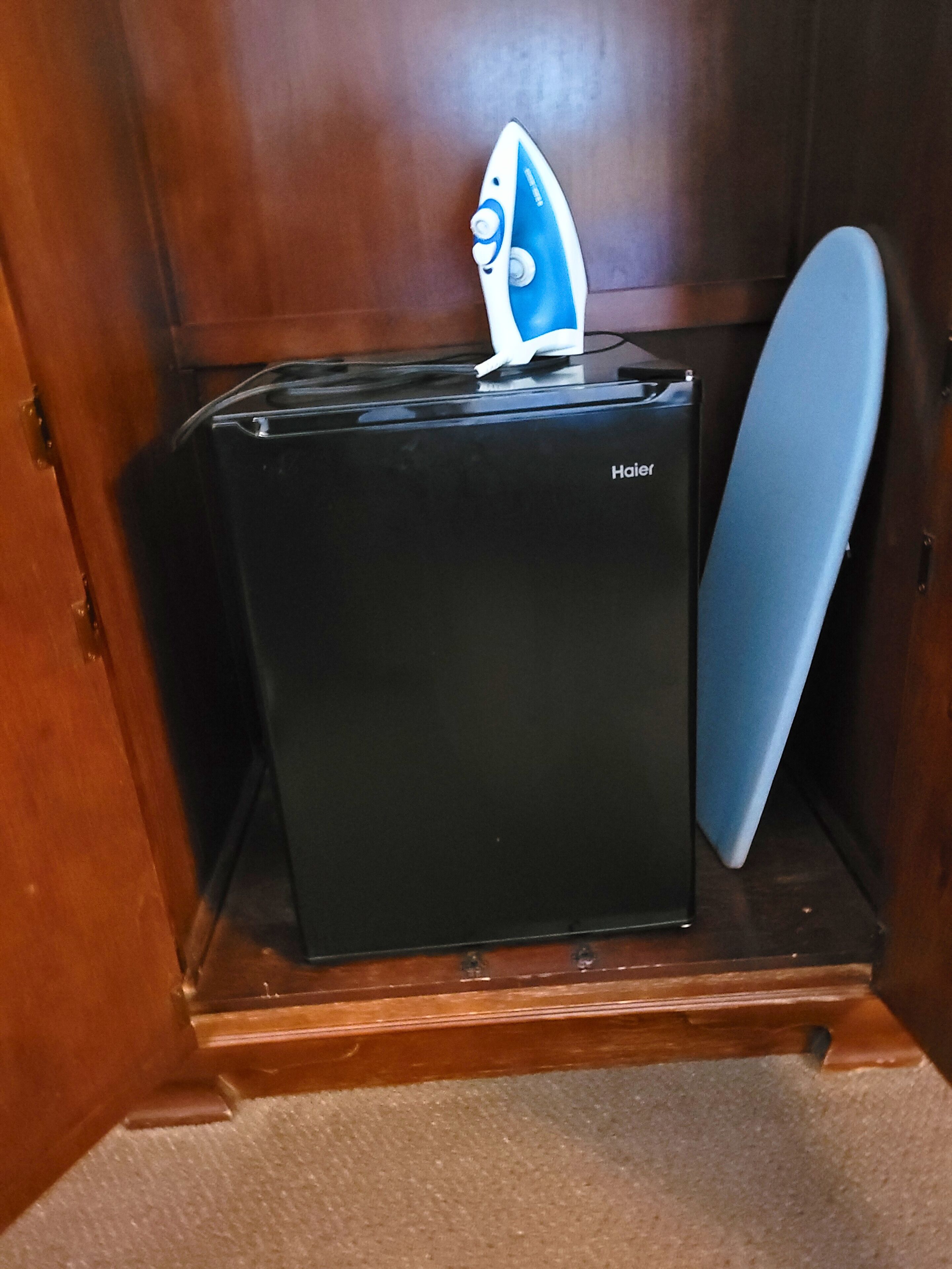 A compact, dark-colored refrigerator with a brand name visible, placed in a wooden cabinet with a blue surfboard leaning against the cabinet.