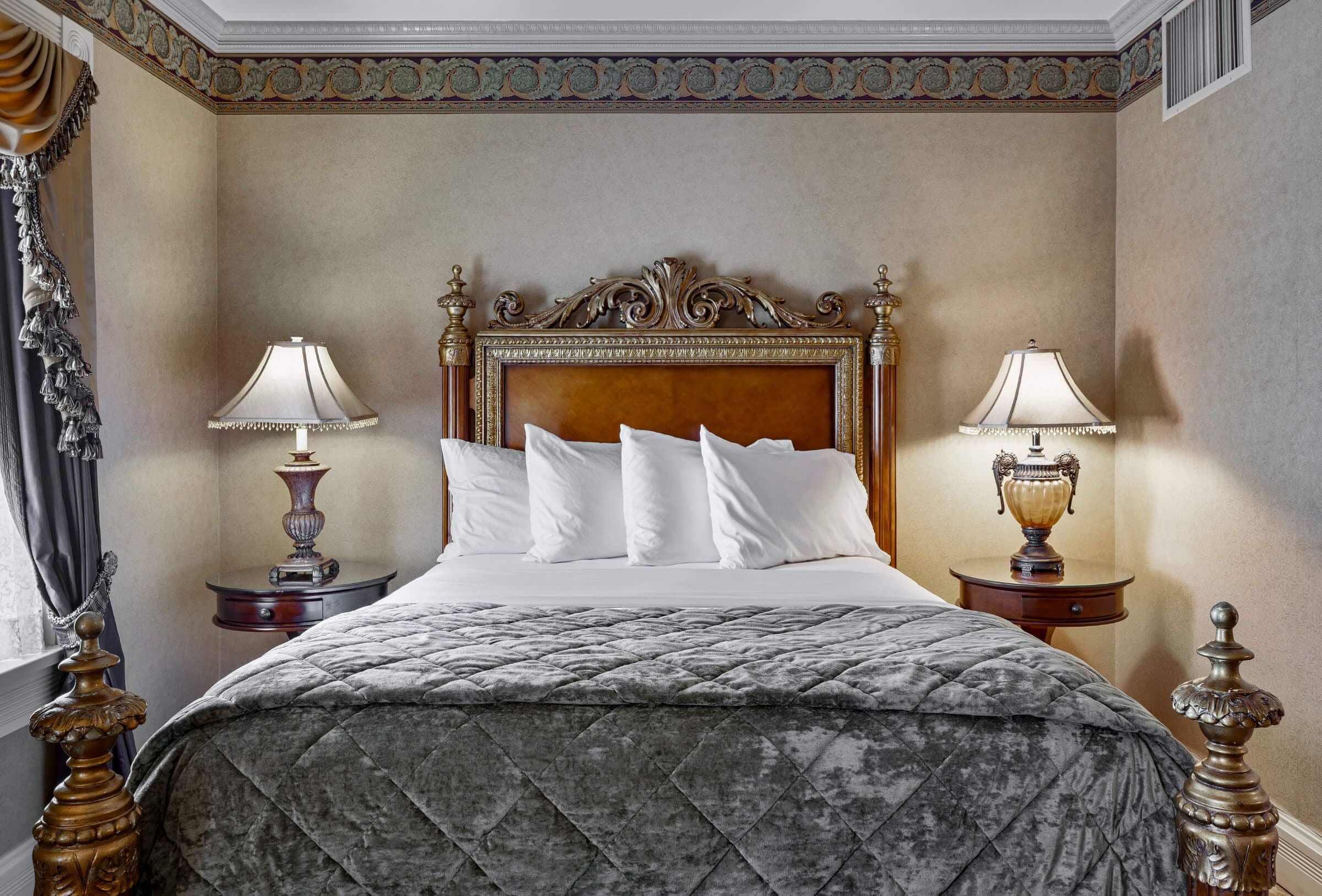Una cama clásica con dosel, con un edredón acolchado, flanqueada por dos mesitas de noche ornamentadas con lámparas, en una habitación con molduras decorativas y una paleta de colores neutros.