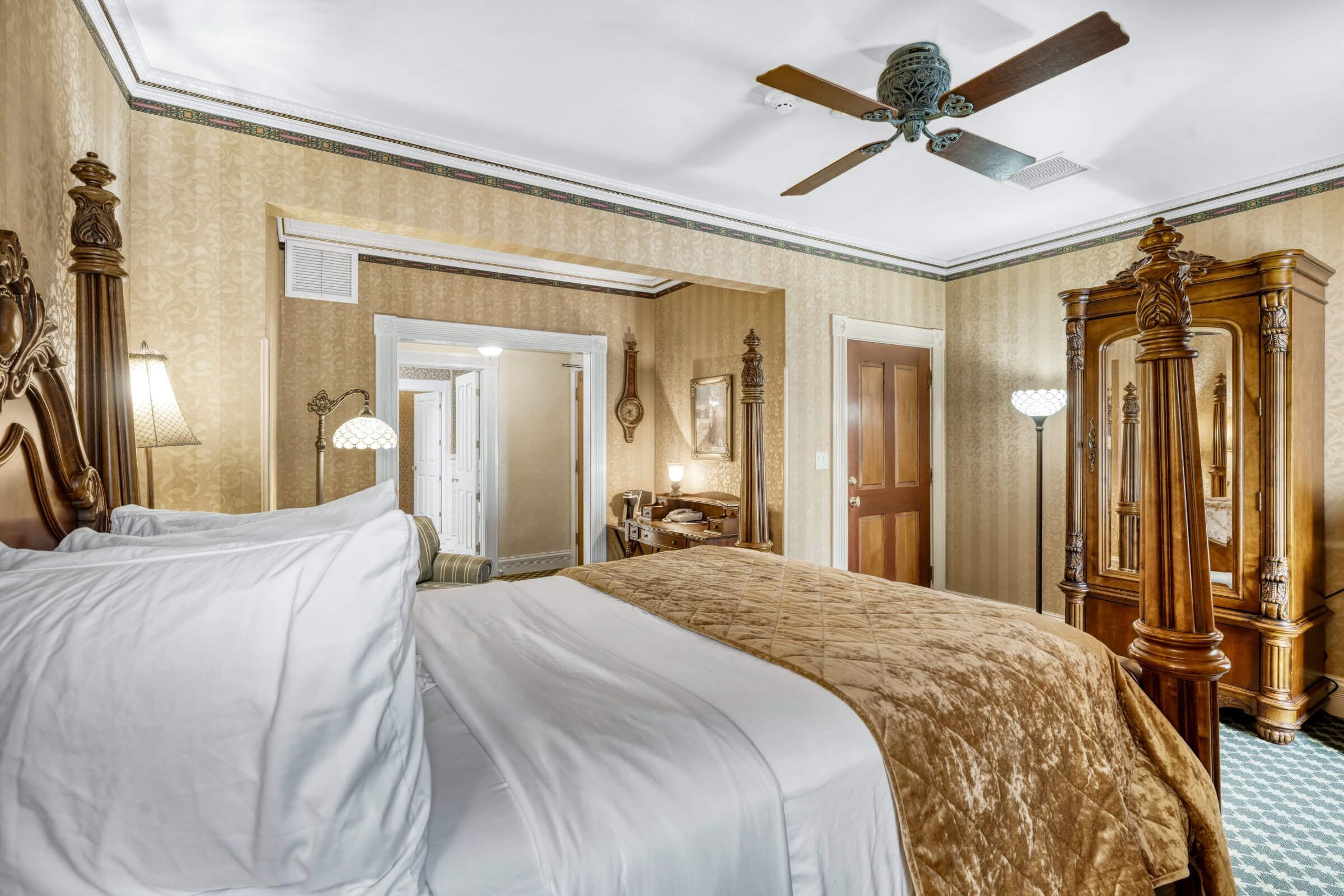 Quarto de hotel com uma cama grande, um ventilador de teto, um guarda-roupa de madeira e uma mesa com abajur.