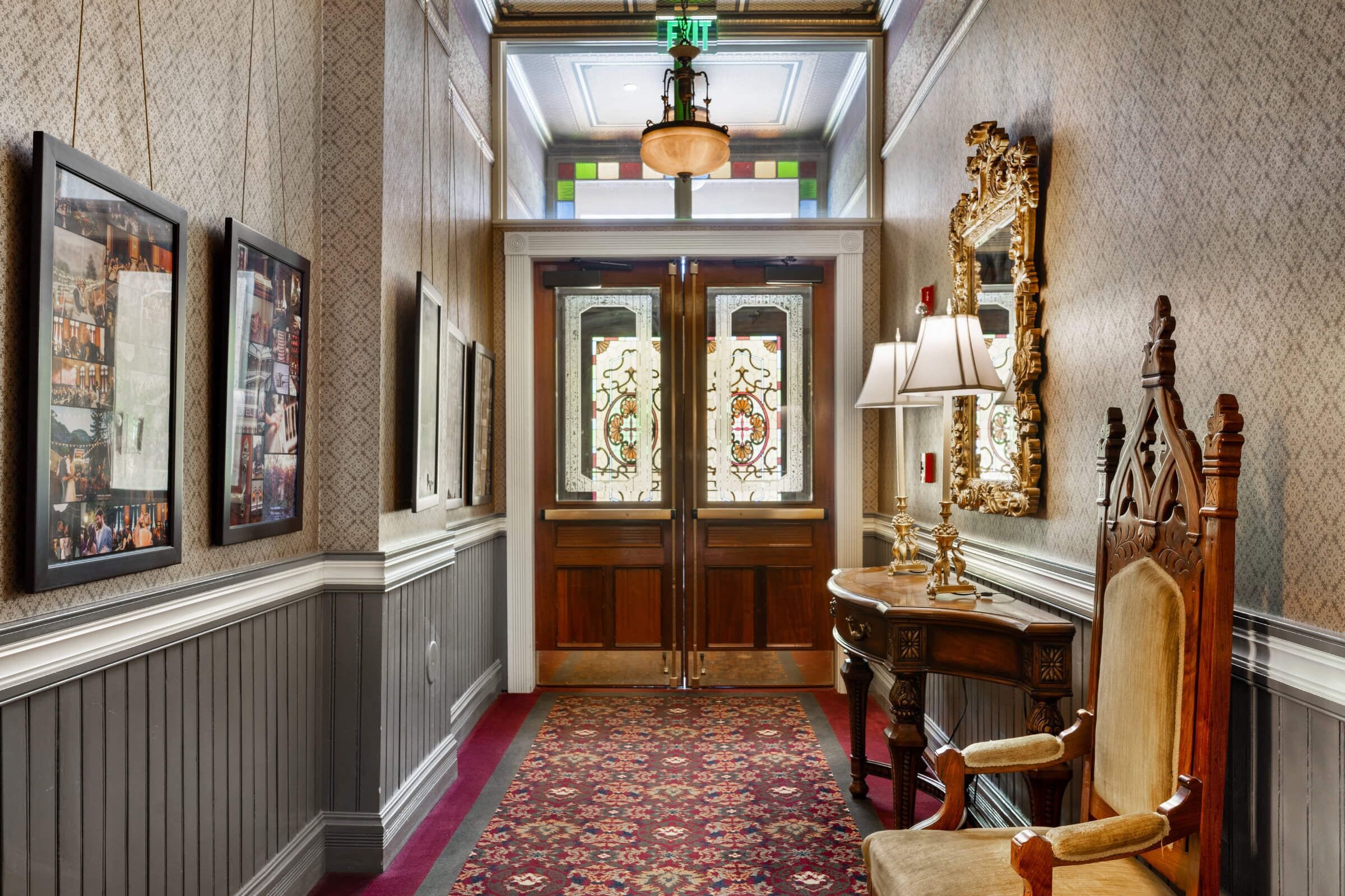 A hallway with a patterned carpet, wooden doors with stained glass, a mirror, and framed pictures on the wall.