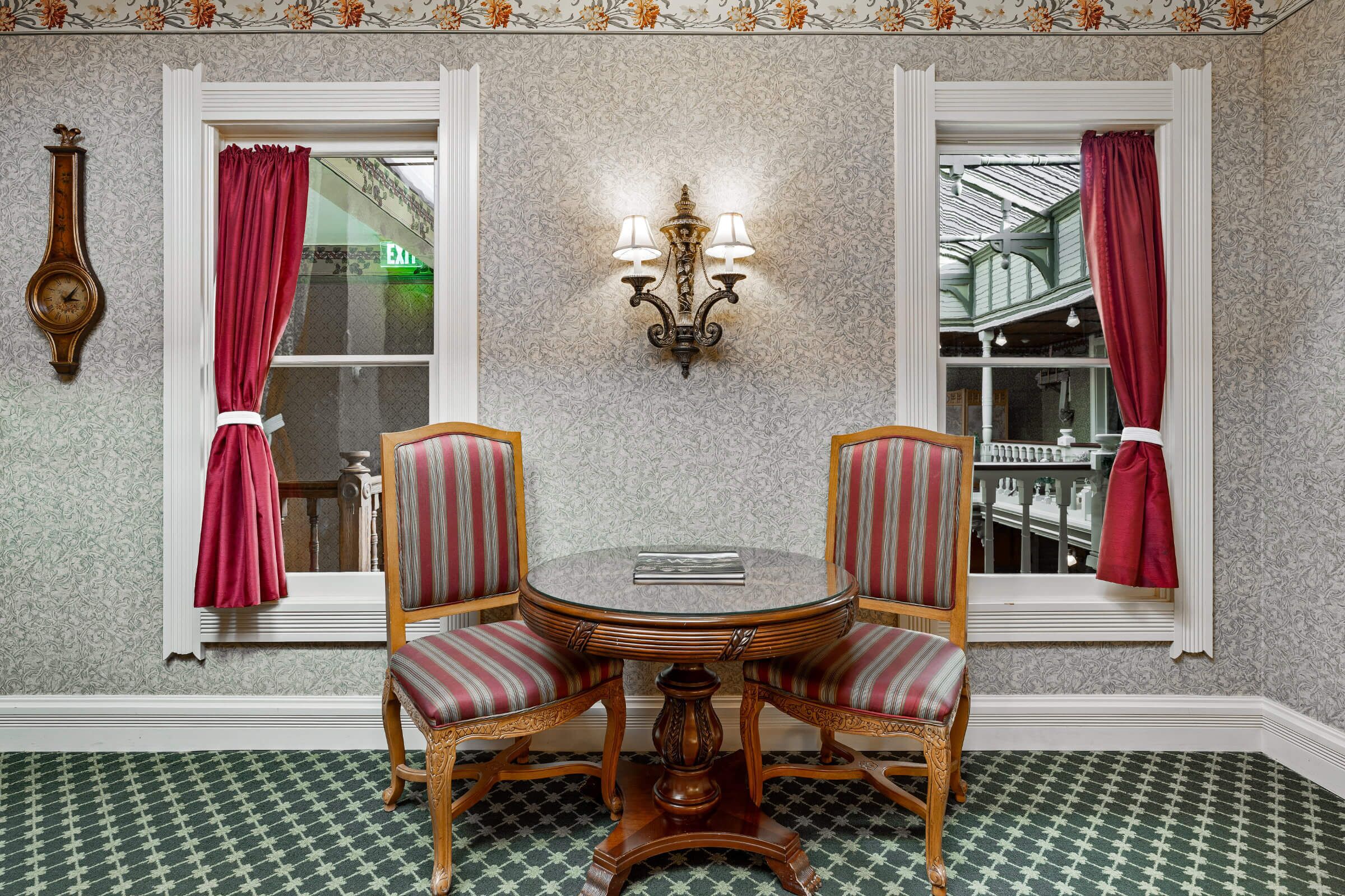 A dining area with a round table, two upholstered chairs, a wall-mounted light fixture, and patterned wallpaper.
