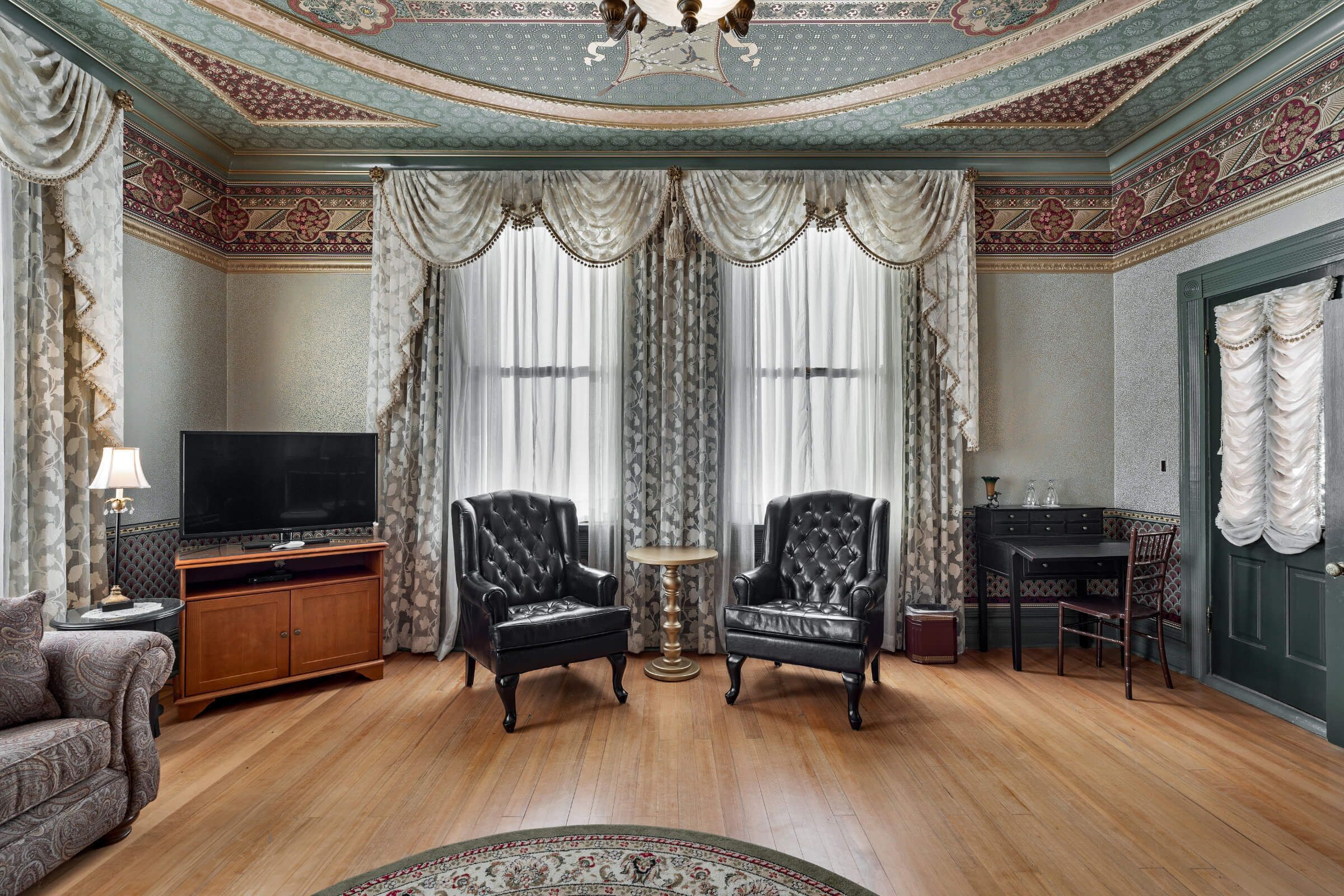 Um quarto com piso de madeira, duas poltronas de couro preto, uma pequena mesa redonda, uma televisão em um móvel de madeira e um tapete estampado.