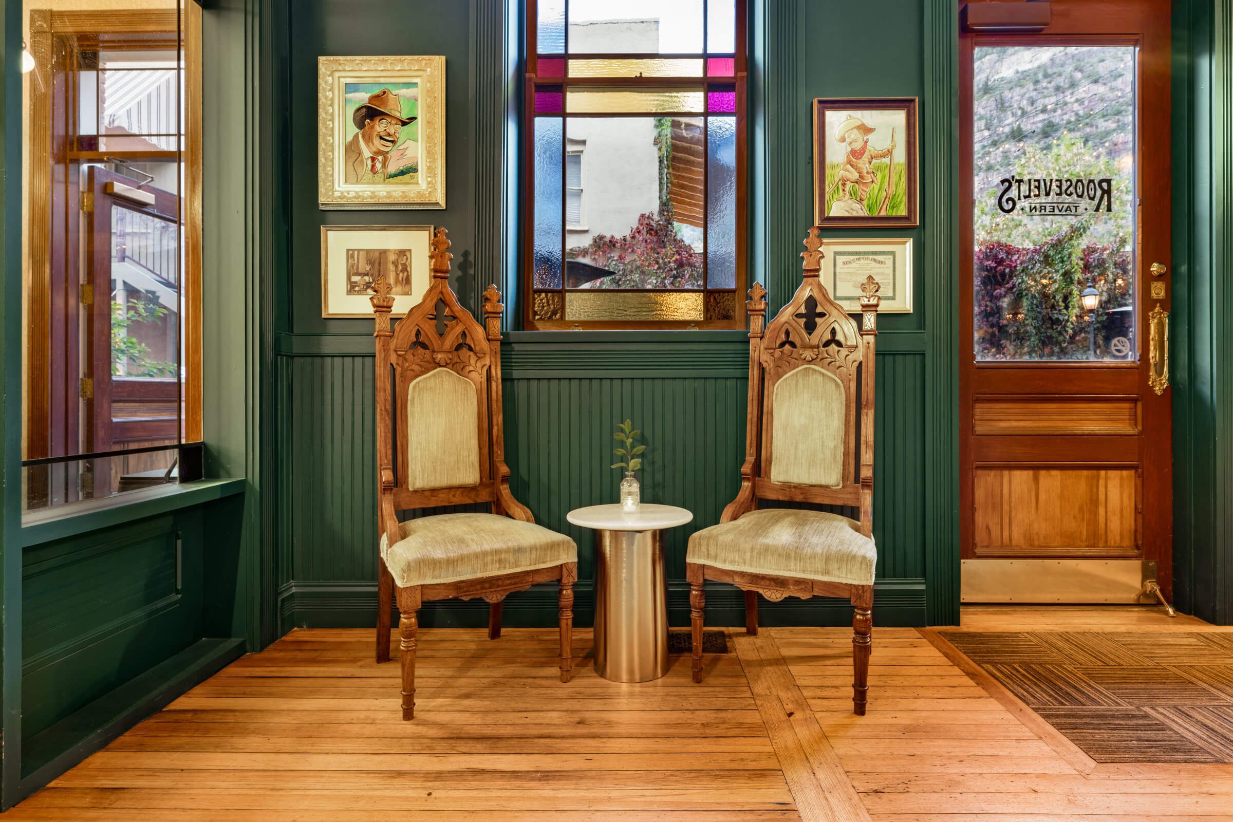A room with two wooden chairs, a small round table, and a mirror above a window with a view of greenery.