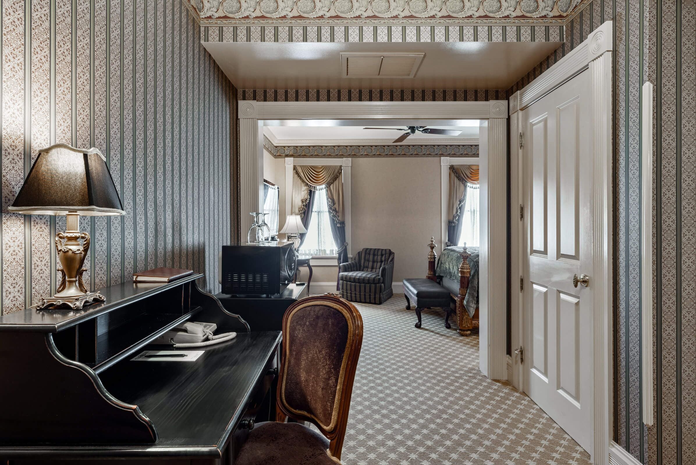 Une pièce comprenant un piano, un bureau avec une lampe, une chaise, un téléviseur et une porte donnant sur une autre pièce avec un lit et une chaise.