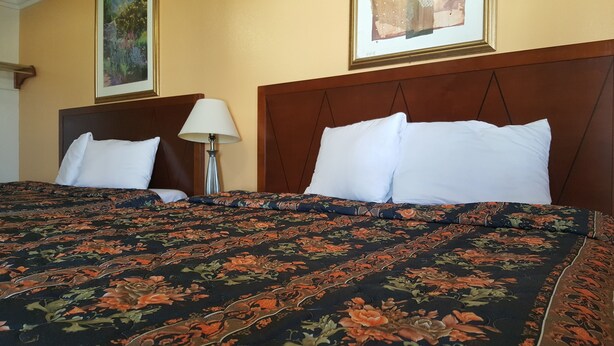 A hotel room with a large bed, two white pillows, a floral-patterned bedspread, a wooden headboard, a bedside lamp, and two framed pictures on the wall. - Royal Inn Motel Long Beach (Long Beach)