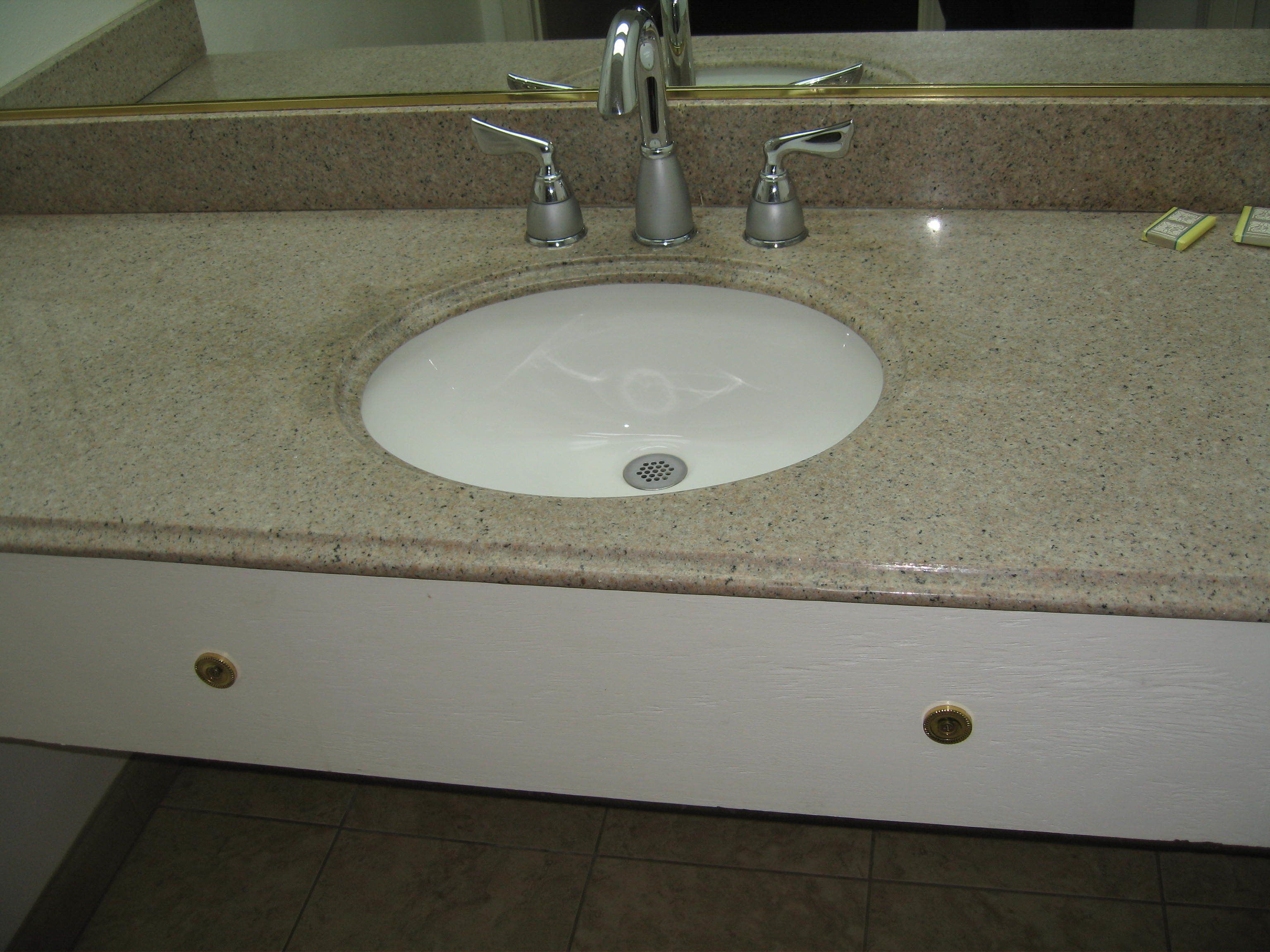 A bathroom sink with a granite countertop, a mirror above it, and two gold-colored faucets.