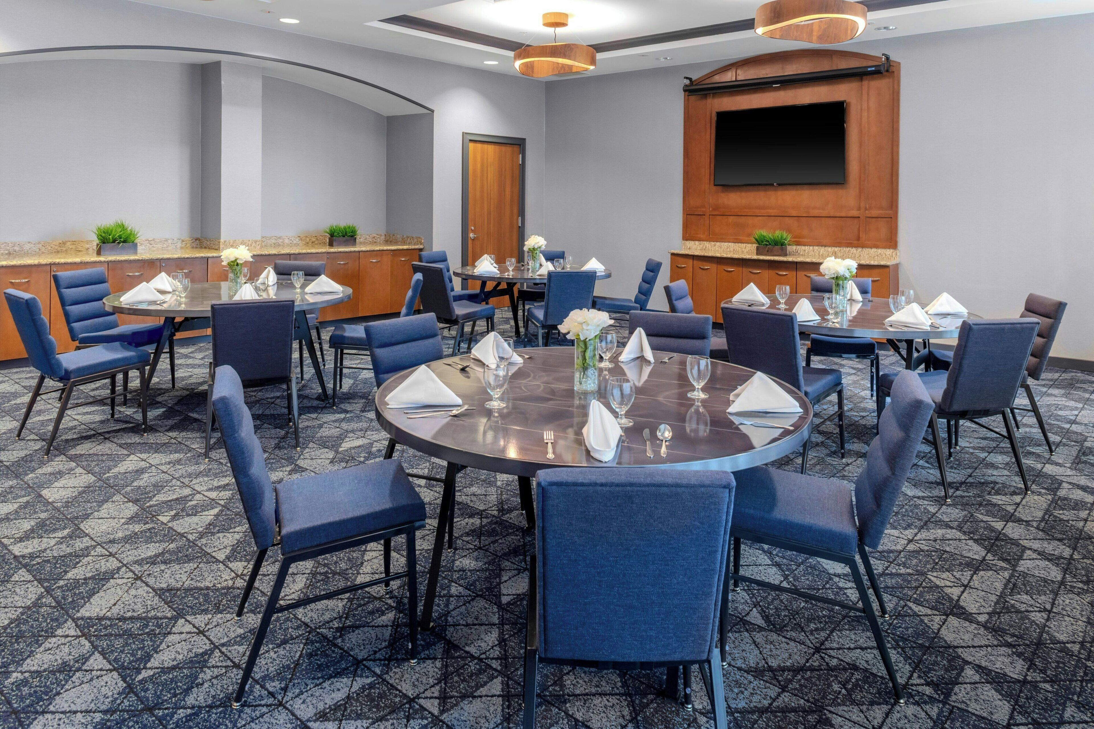 A conference room with round tables, blue chairs, and a large flat-screen TV on the wall.