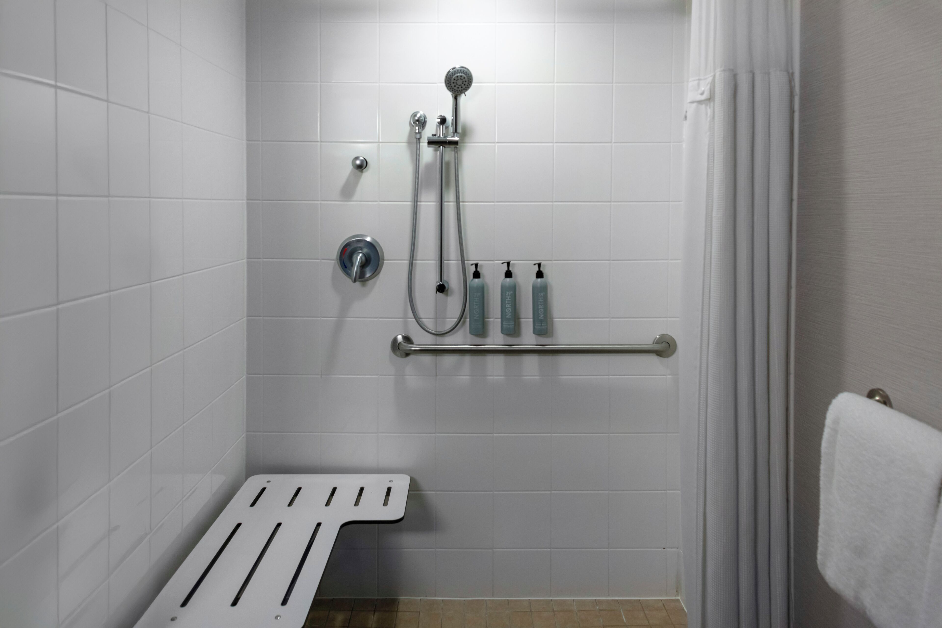 A small, white-tiled bathroom with a shower, a towel rack, and a handrail.