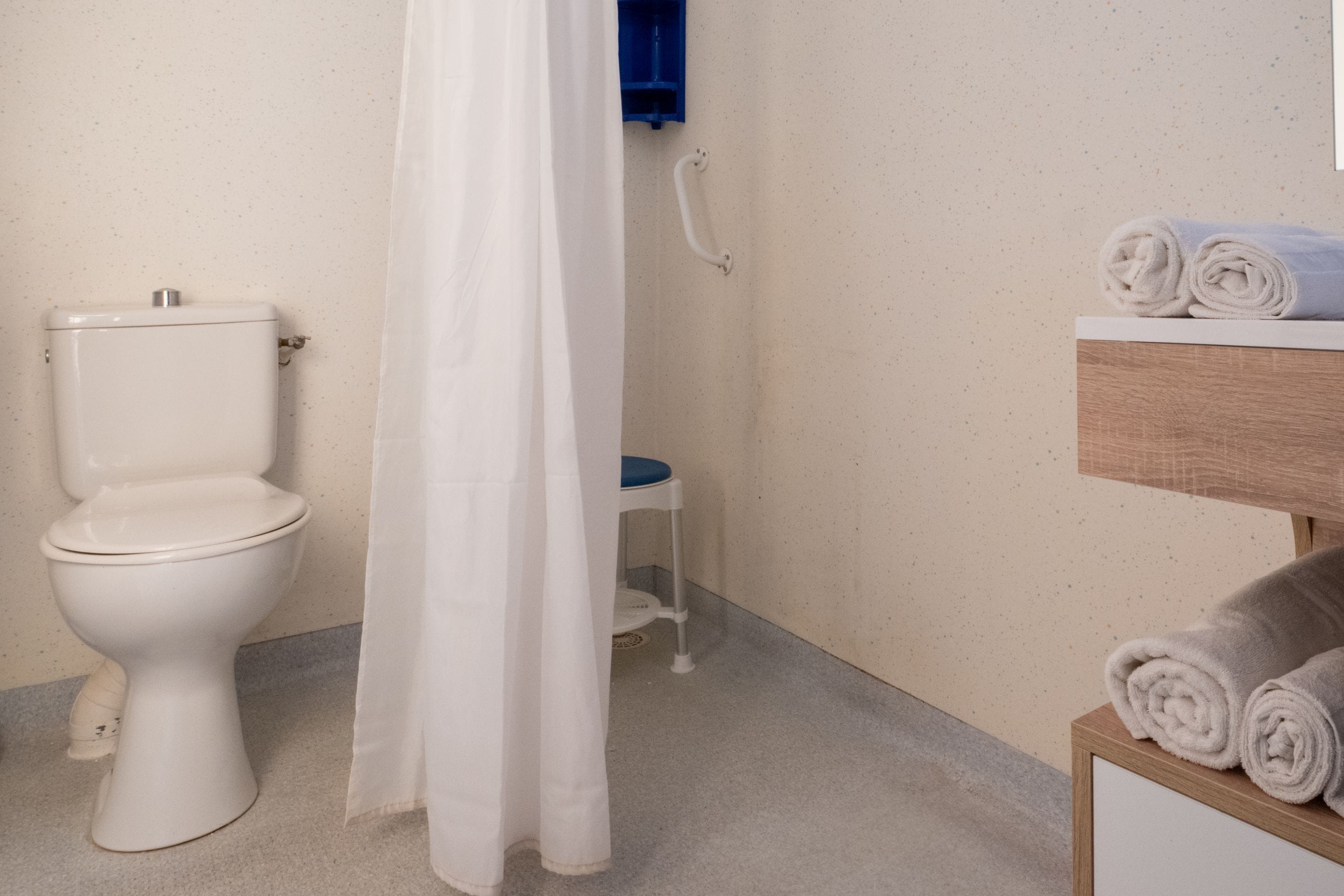 Ein Badezimmer mit Toilette, einer Duschvorhang, einem kleinen Hocker und einem Holzregal mit zusammengerollten Handtüchern.