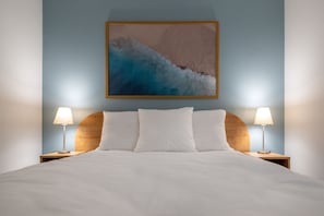 Un lit bien fait, avec du linge de lit blanc, deux lampes de chevet et une œuvre d’art encadrée sur le thème de la côte, accrochée au mur.