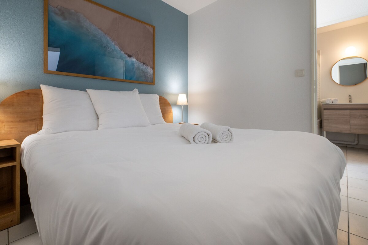 A neatly made bed with white linens and two rolled towels, a wooden headboard, a bedside table with a lamp, and a painting on the wall.