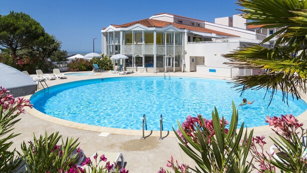 A swimming pool with lounge chairs and umbrellas, surrounded by a building with a tiled roof and a view of the sea. - Résidence Odalys Les Terrasses de Fort Boyard (Fouras)
