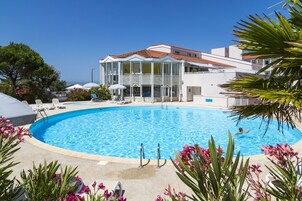 Une piscine entourée de chaises longues et de parasols, bordée d’un bâtiment doté d’un toit en tuiles et offrant une vue sur la mer.