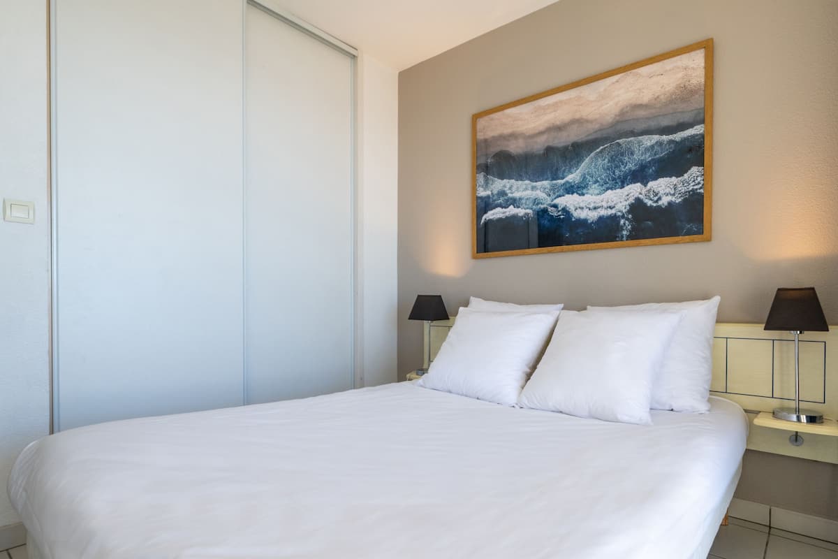 Une chambre d’hôtel avec un grand lit, deux tables de chevet avec des lampes, un tableau représentant des vagues accroché au mur et une porte fermée.