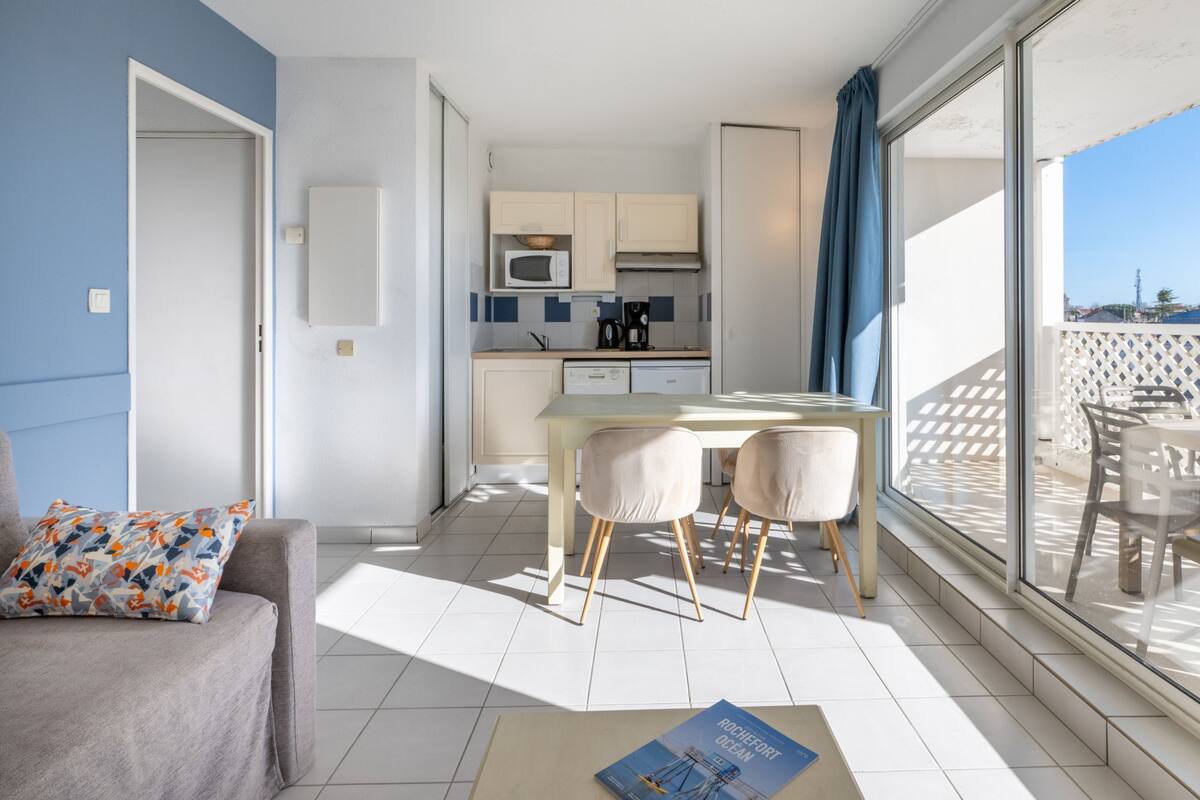 Un appartement moderne doté d’une kitchenette, d’un coin repas avec deux chaises, d’un canapé agrémenté d’un coussin à motifs et d’un balcon offrant une vue agréable.
