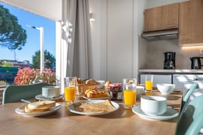 Une table de petit-déjeuner avec des assiettes de toasts, des croissants et du jus, une machine à café, et une vue sur l’extérieur.