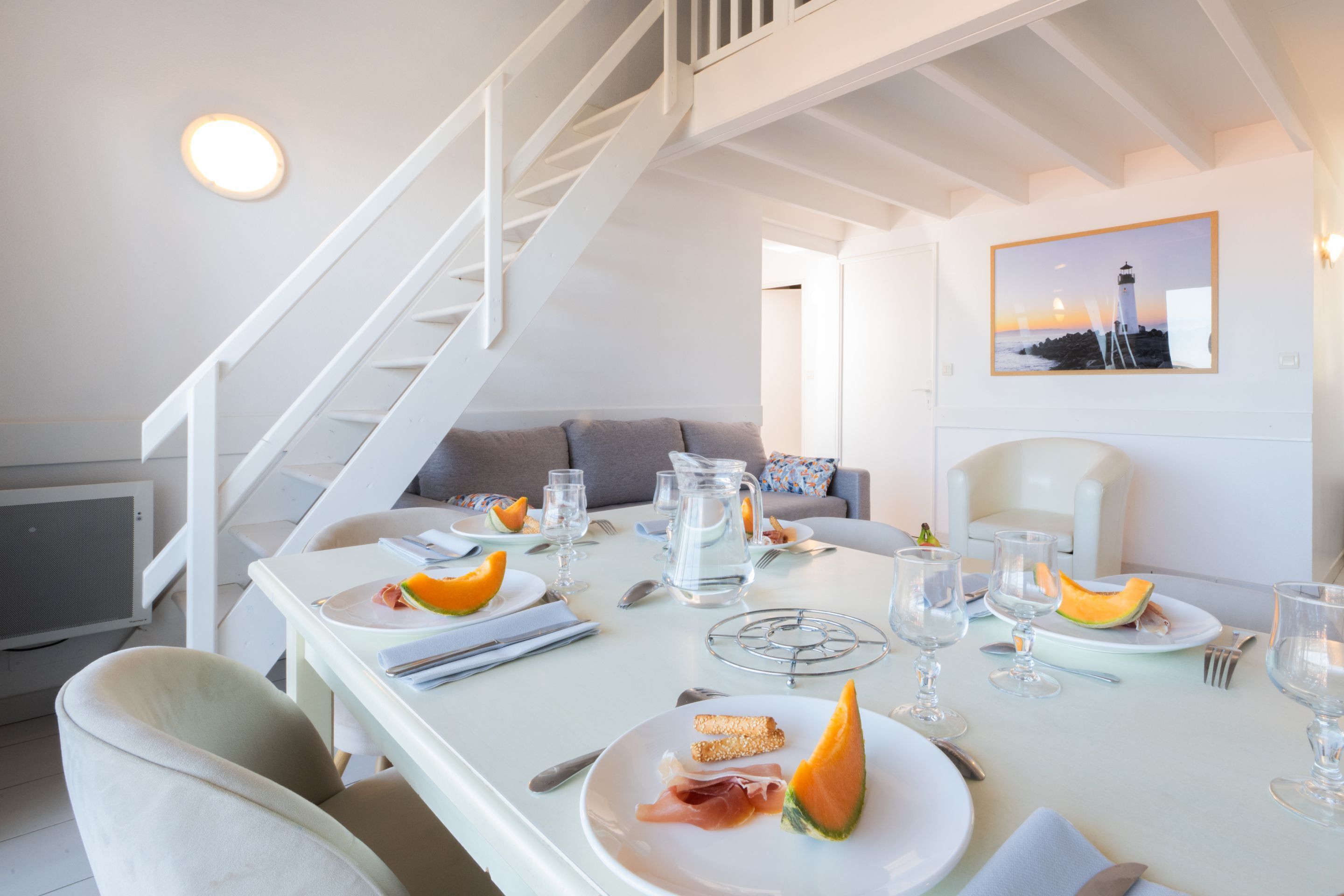 Un comedor con una mesa blanca dispuesta para dos personas, con platos que incluyen comida y bebidas, una jarra de agua y un cuadro de un faro en la pared.