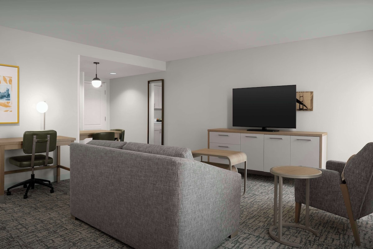 Une chambre d’hôtel moderne avec un canapé gris, une petite table ronde, un téléviseur posé sur un meuble et un bureau avec une chaise.