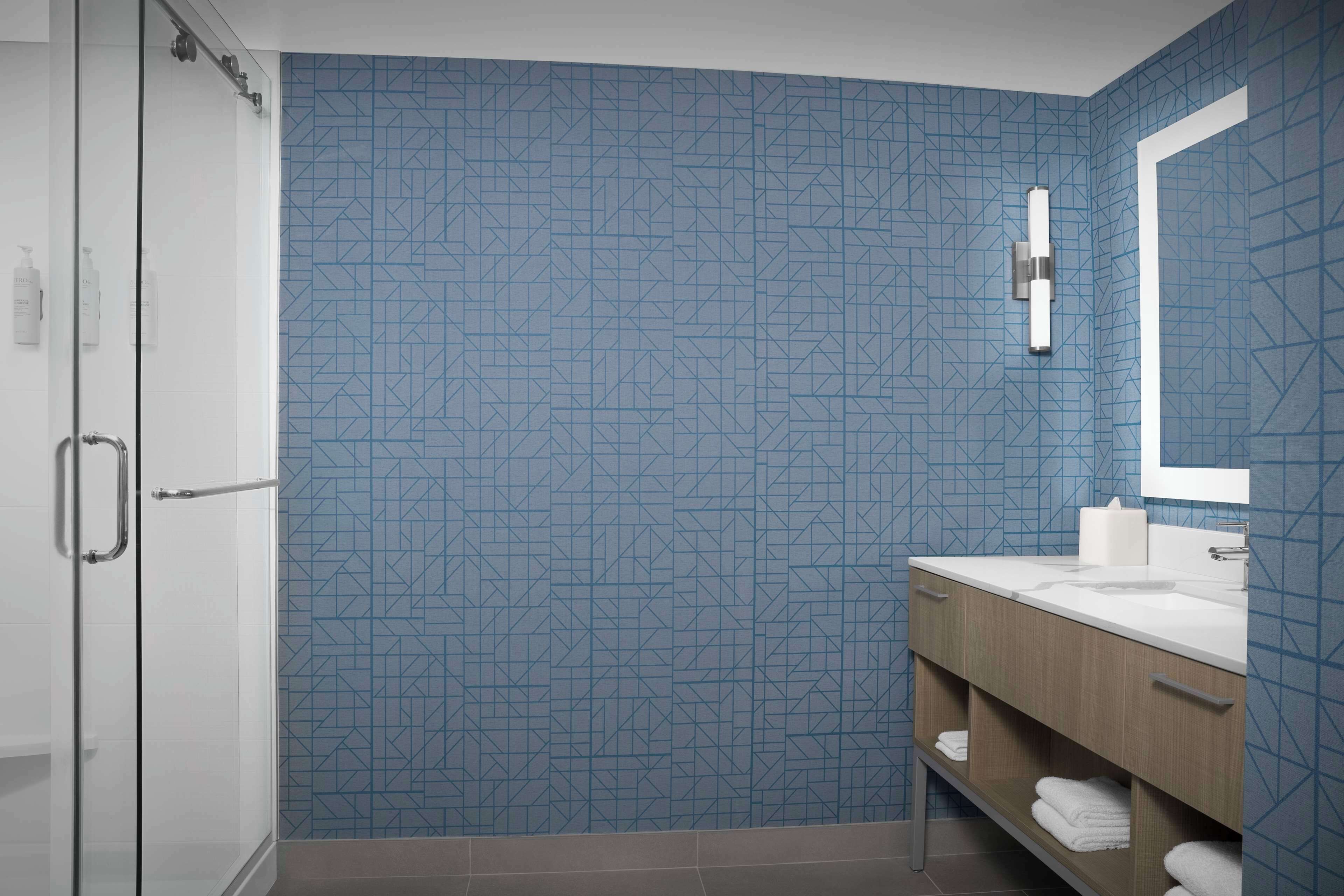 Een moderne badkamer met een geometrisch tegelpatroon, een glazen douchecabine, een houten wastafelmeubel met een witte wasbak en een grote spiegel.